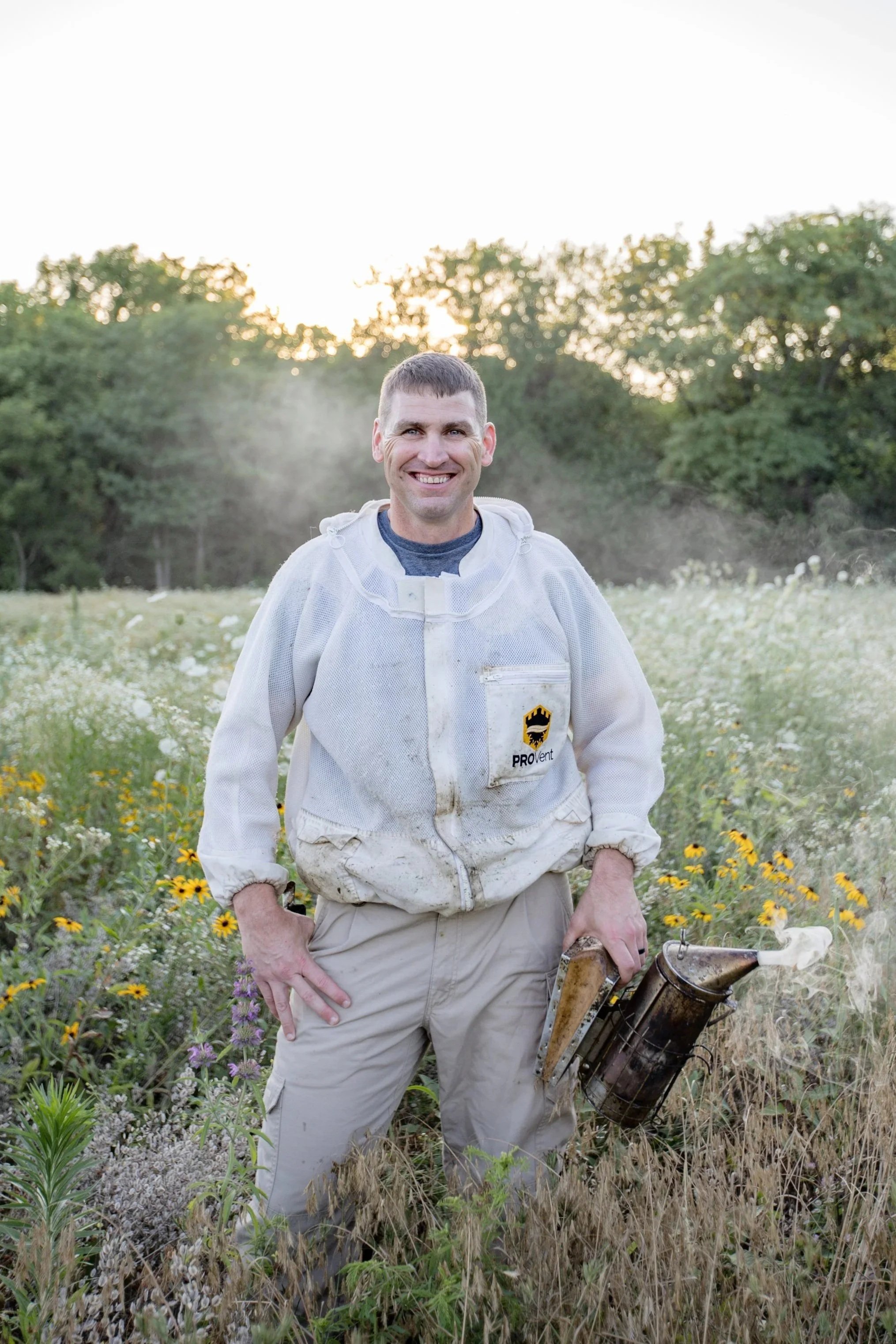 A man in a beekeeping suit standing in a field of wildflowers with trees in the background, holding a smoker used for bee management.