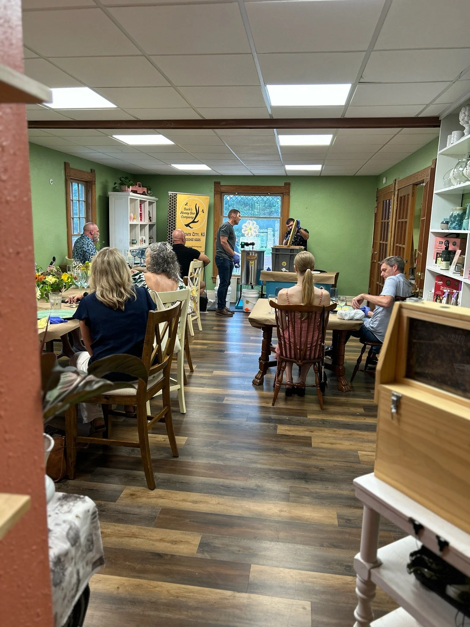 A group of people gathered in a cozy, well-lit room with green walls, wooden trim, and wooden floors, listening to a musician performing on a small stage. The audience is seated at tables and enjoying the event, with a colorful banner and shelves filled with decorative items in the background.