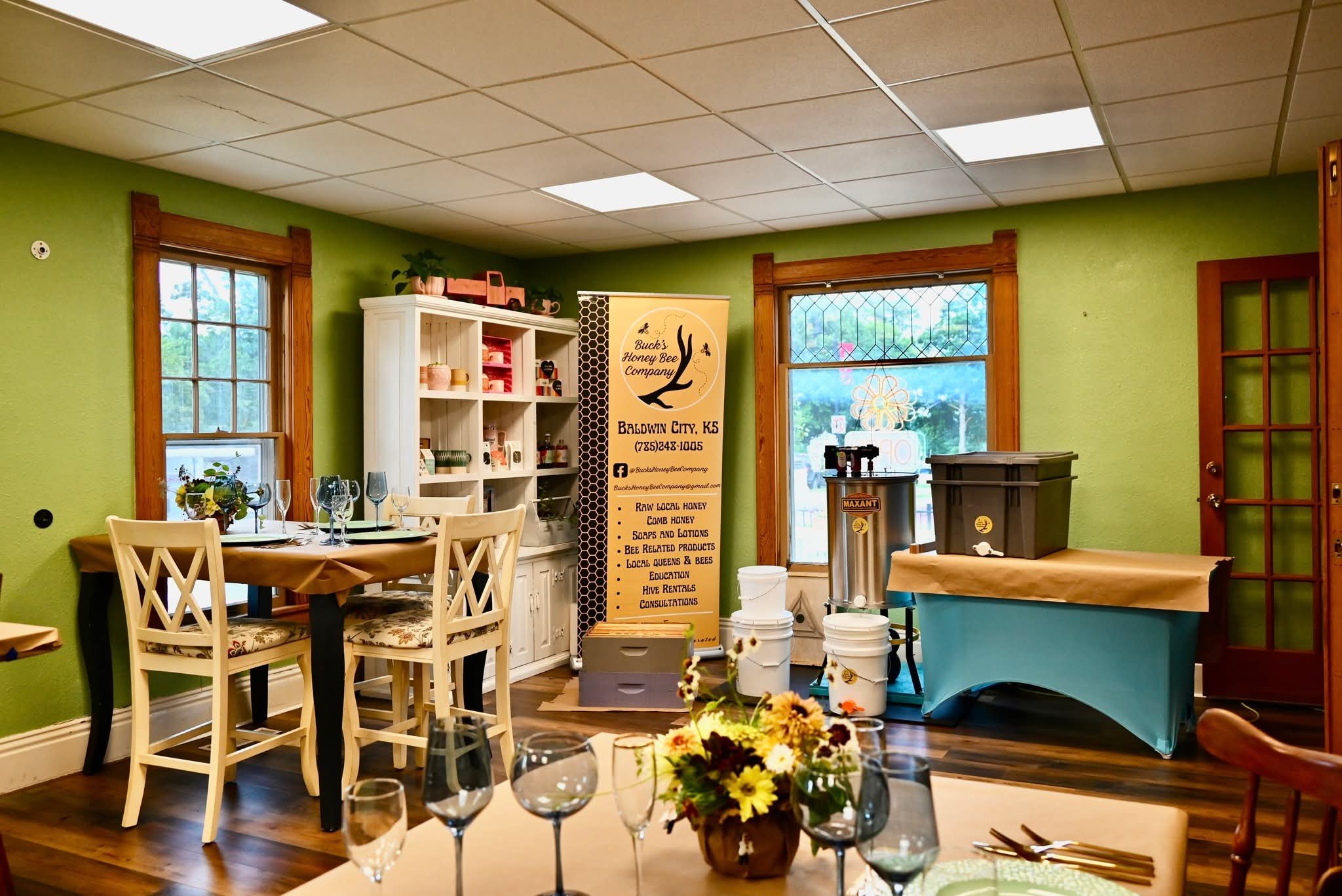 Interior of a room with green walls, wooden trim, a dining table set with glassware and a flower arrangement, a white cabinet filled with jars and supplies, a banner for Buck's Honey Bee Company, and event setup equipment like buckets and storage containers.