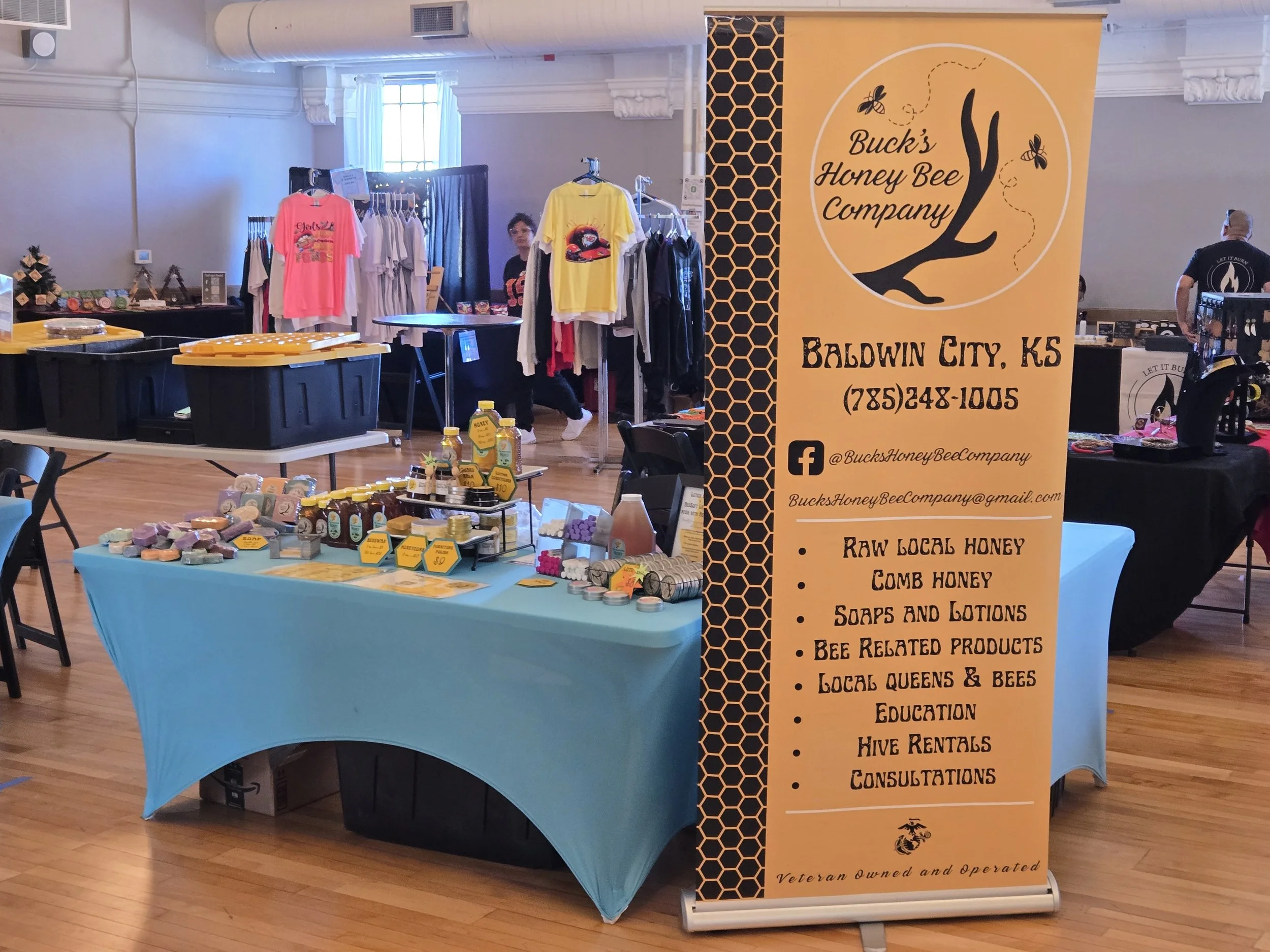 A vendor booth for Buck's Honey Bee Company set up at an indoor market, featuring honey products, soaps, and lotions, with a bright orange banner displaying contact information and product offerings.