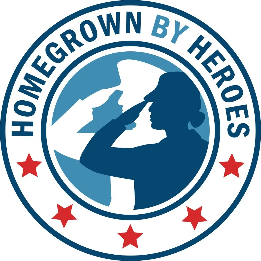 A circular logo with a woman in profile saluting, holding a flag, with the American map outline in the background. The text around the circle reads "Home Grown By Heroes" and there are red stars at the bottom.