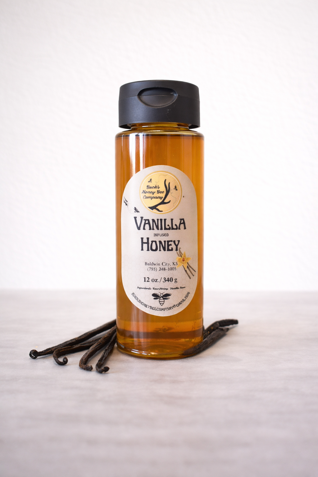 Amber bottle of vanilla infused honey with a black lid, placed on a light surface with vanilla beans around its base.
