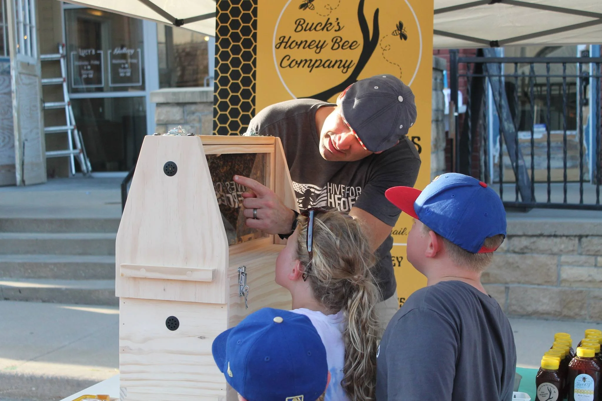 A man wearing a black shirt and a dark baseball cap demonstrating honeybees to children at a booth with a sign that reads 'Buck's Honey Bee Company.' The children are wearing caps, with one girl having sunglasses on her head. Honey jars are on the table.