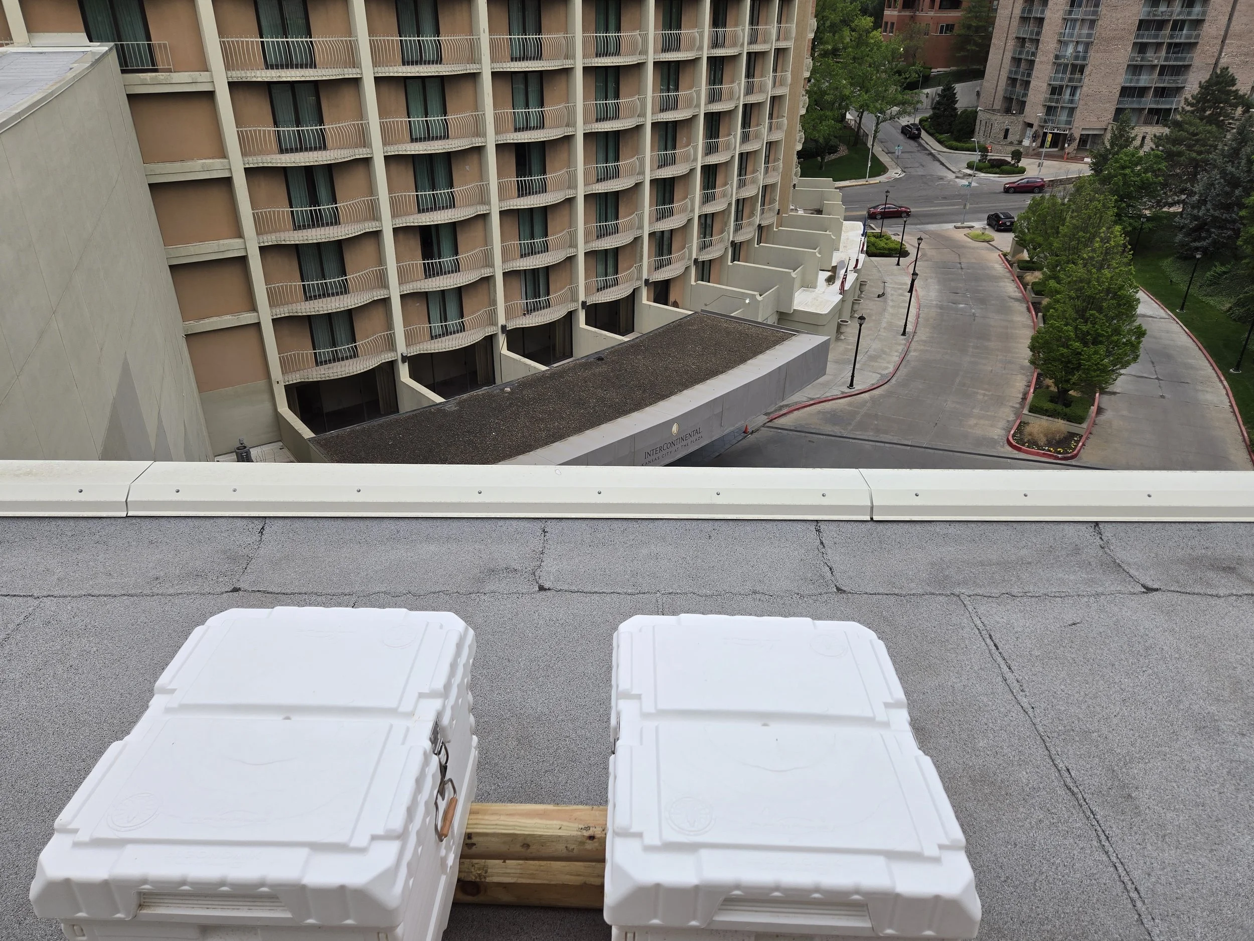 View from a rooftop parking lot with two white insulated coolers placed on wooden planks, looking over a city street with trees and apartment buildings.
