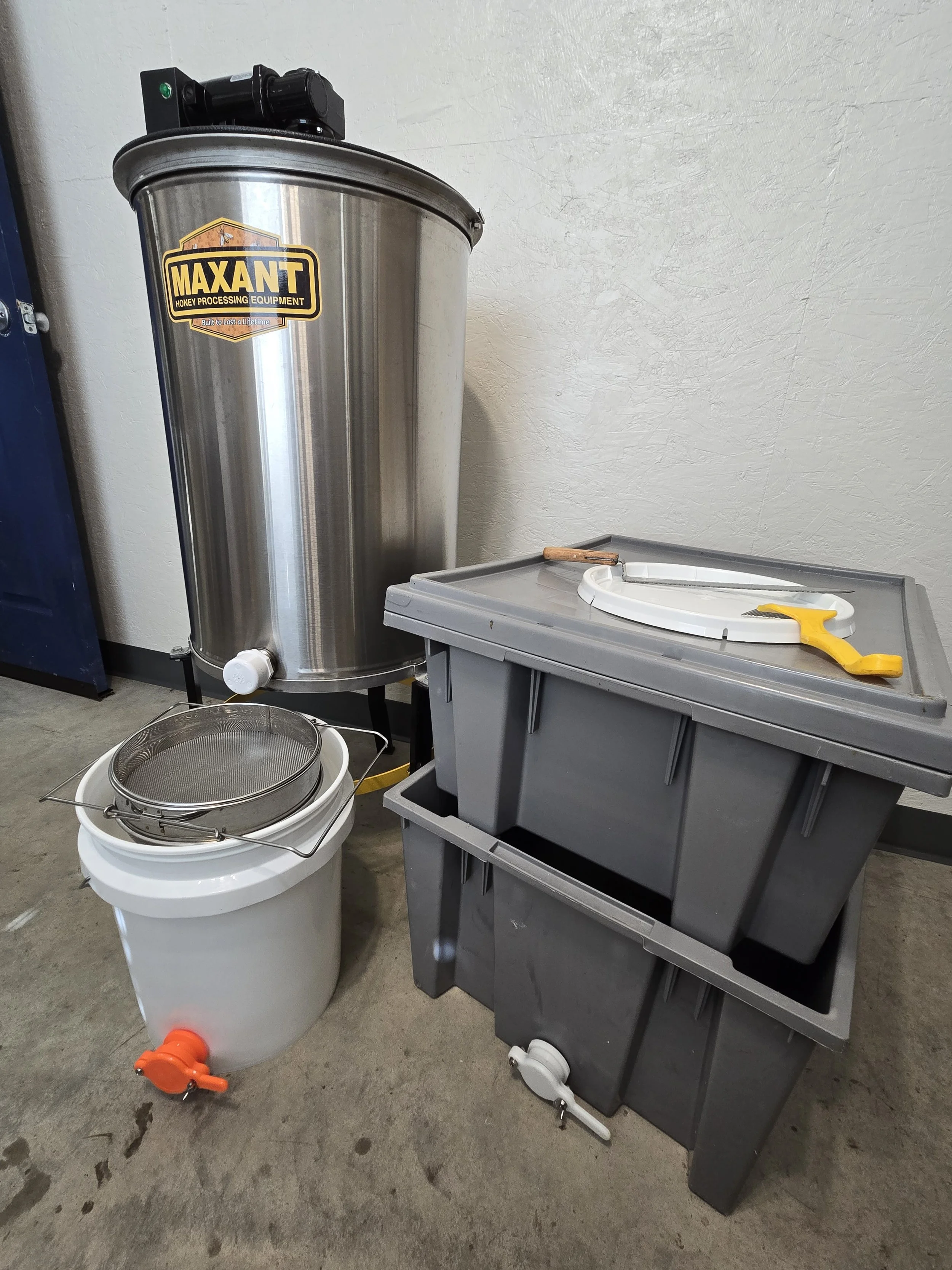 Large stainless steel honey processing equipment with the brand Maxant, two gray plastic tanks with lids, a white bucket with a strainer on top, and honey processing tools including a scraper and a saw, all placed on a concrete floor against a white wall.