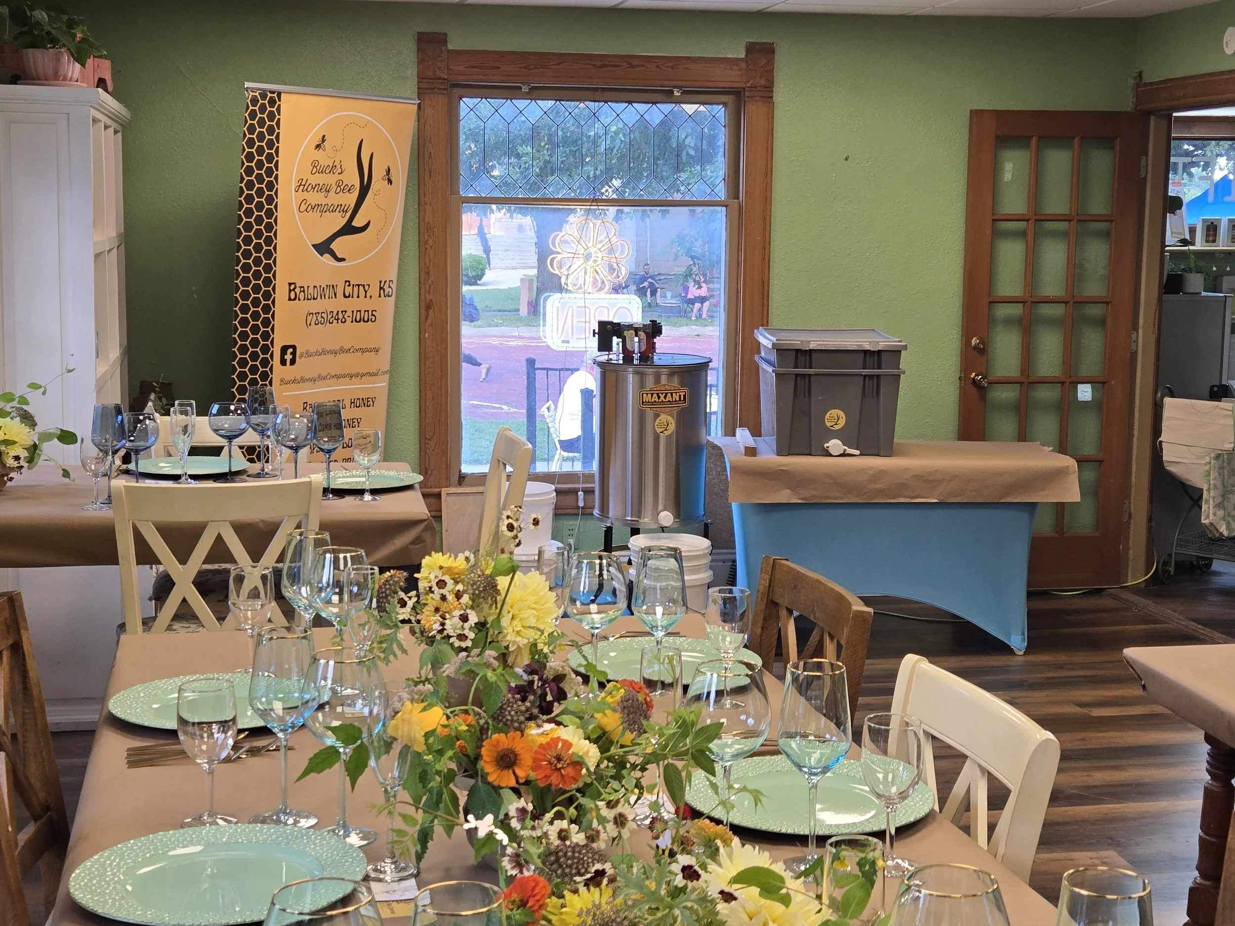 Indoor dining area set for a celebration with banquet tables, floral centerpieces, glassware, and a beverage station with a honey warmer. Green walls, wooden trim, large window, and a door are visible.