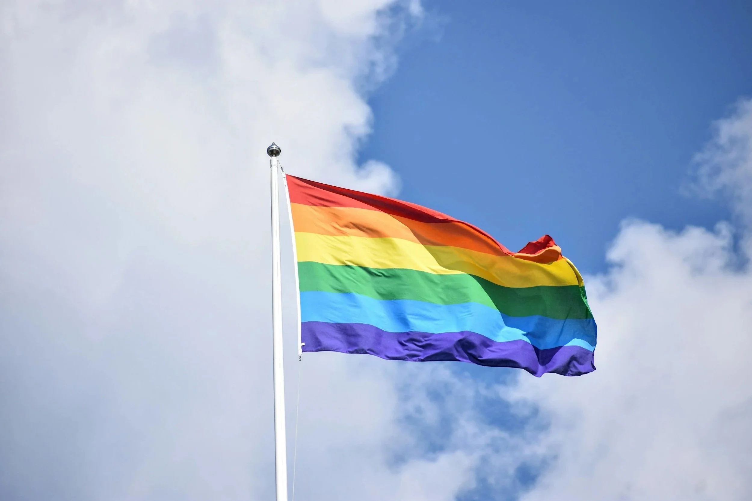 A rainbow Pride flag waving in the sky with white clouds.