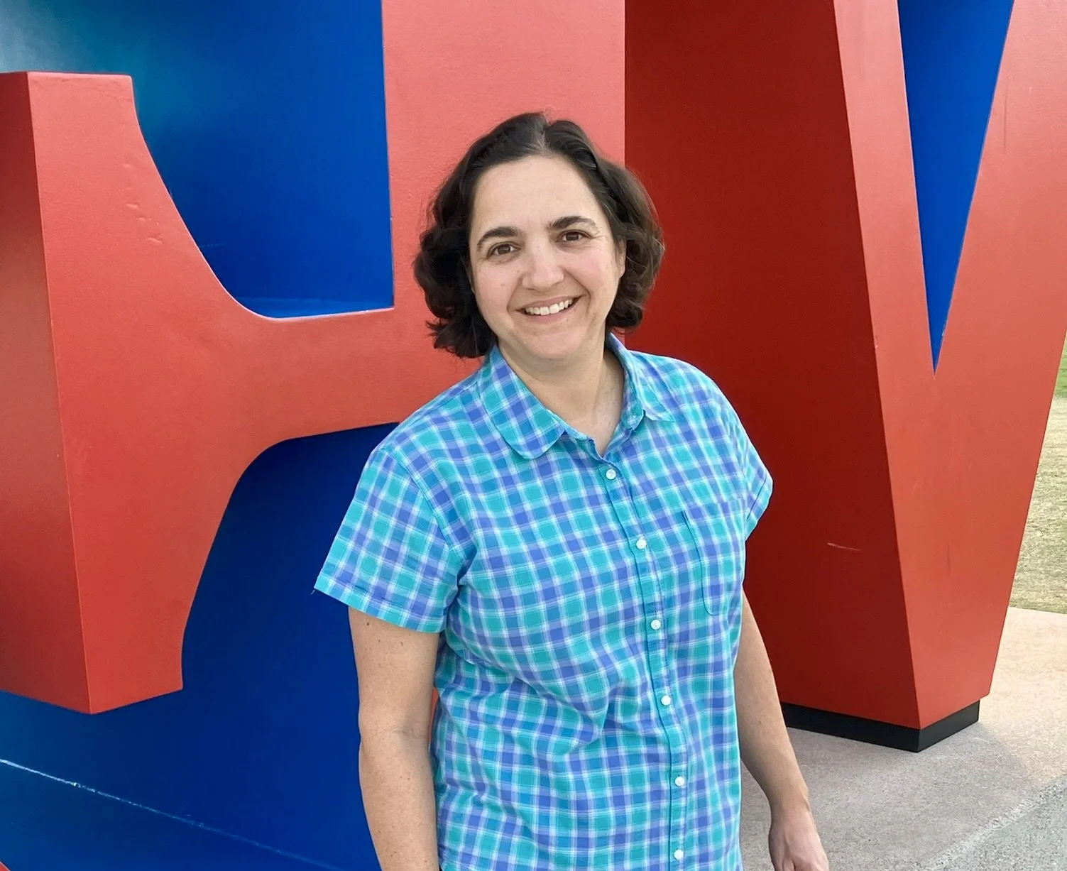 A woman with short dark hair wearing a blue and green plaid shirt, standing outdoors beside large pop art-style letters in red and blue.