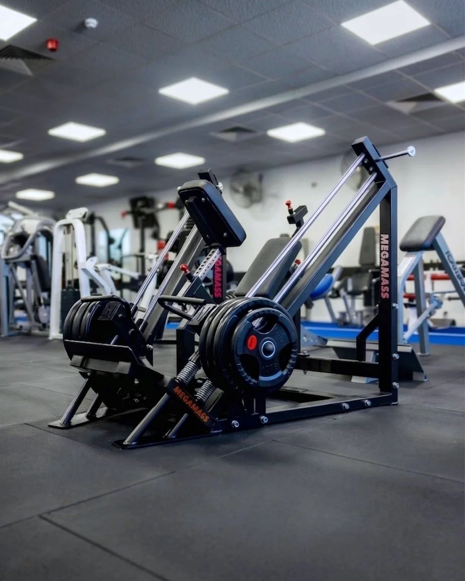 MEGAMASS back training machine in foreground, surrounded by various gym equipment, bright white ceiling lights, and a black rubber gym floor. At Our Gym Fitness Club (Burnside)