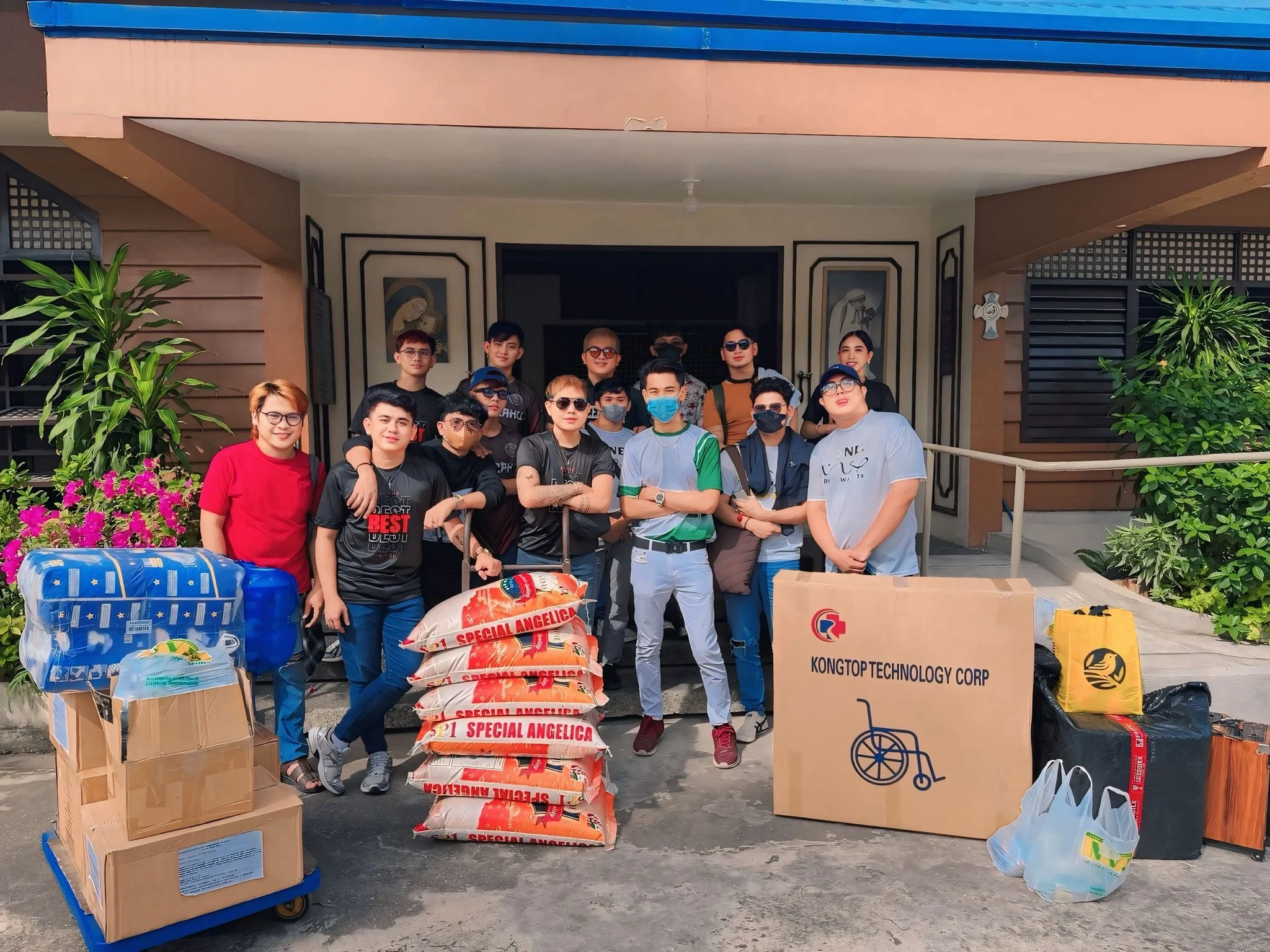 A group of people standing in front of a building with packages, bags, and boxes on the ground, including a large box with "KONGTOP TECHNOLOGY CORP" and a wheelchair icon, and bags labeled "Special Angelica."
