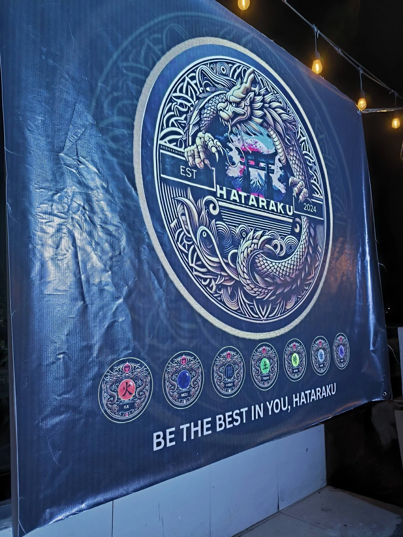 A large, circular emblem featuring a stylized dragon and Japanese elements, with the text 'HATARAKU 2024' and 'EST' on either side. Below the emblem are seven smaller symbols with Japanese characters, and the phrase 'BE THE BEST IN YOU, HATARAKU.'