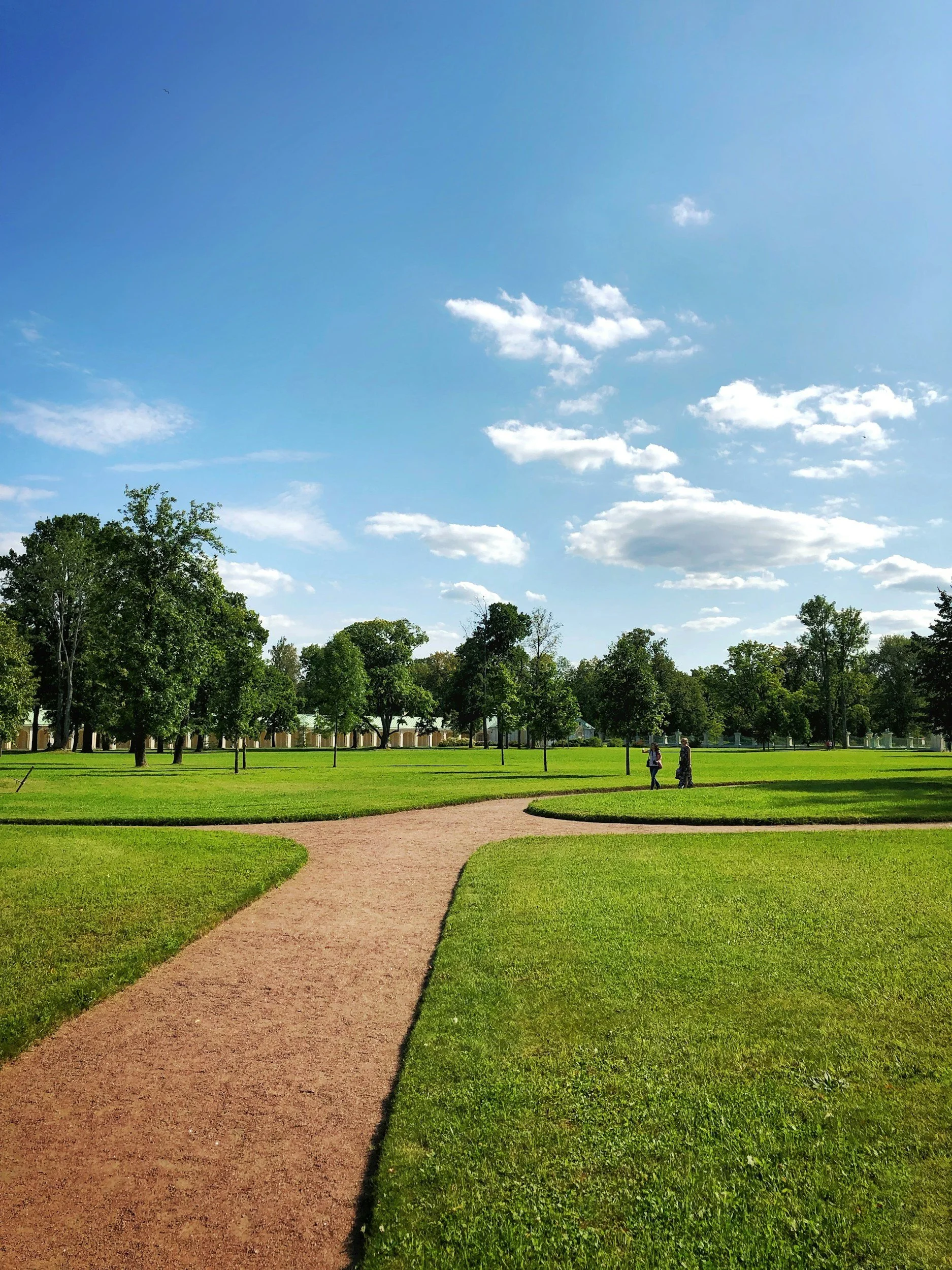 A park with a dirt path winding through lush green grass, trees in the background, and a partly cloudy blue sky.