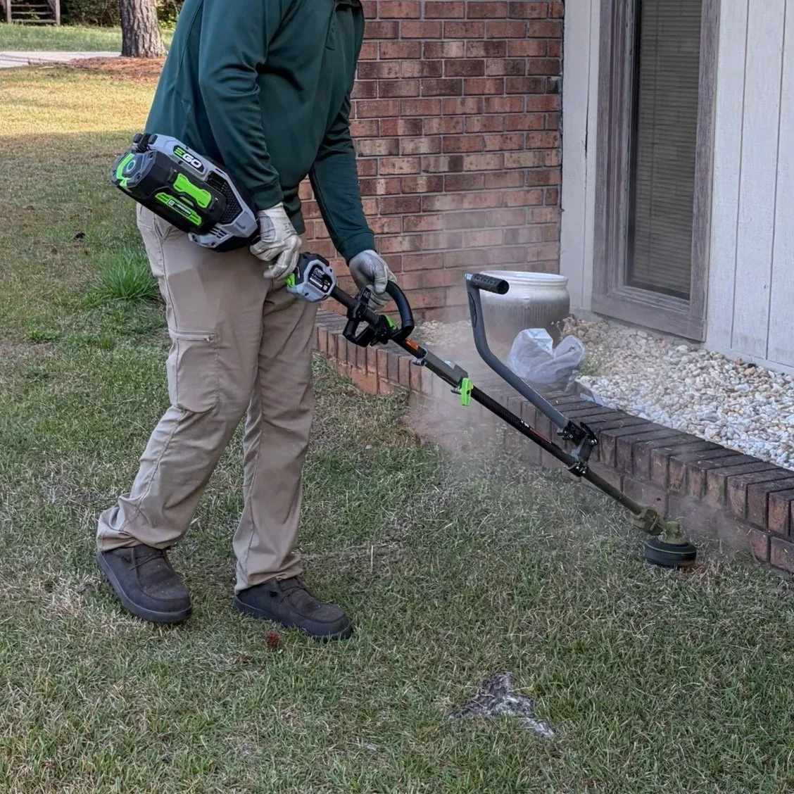 A person using a cordless electric weed trimmer to cut grass near a brick house and window, with a grassy yard in the background.