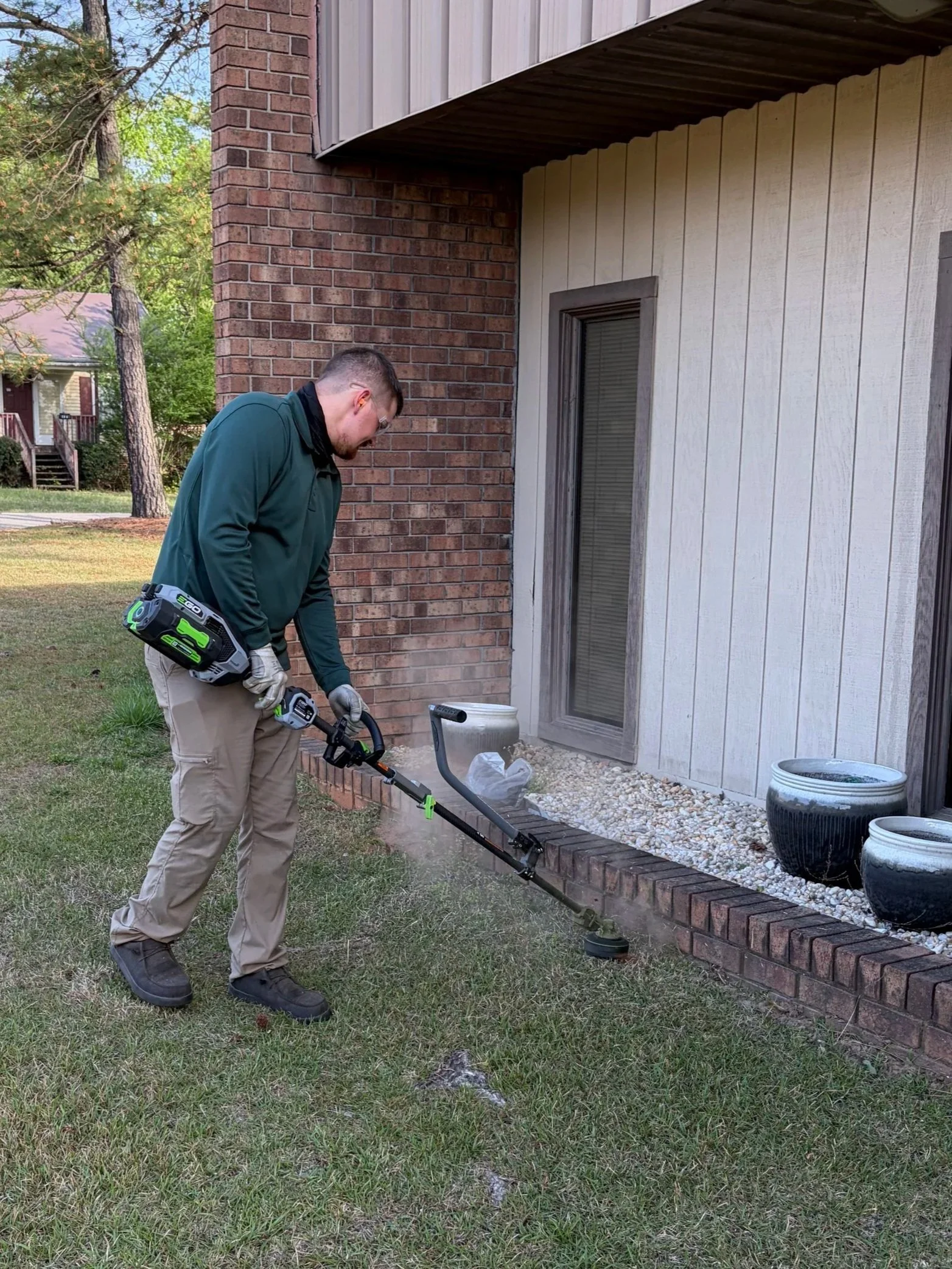 A man operating a metal detector outdoors near a house with brick and wooden siding.