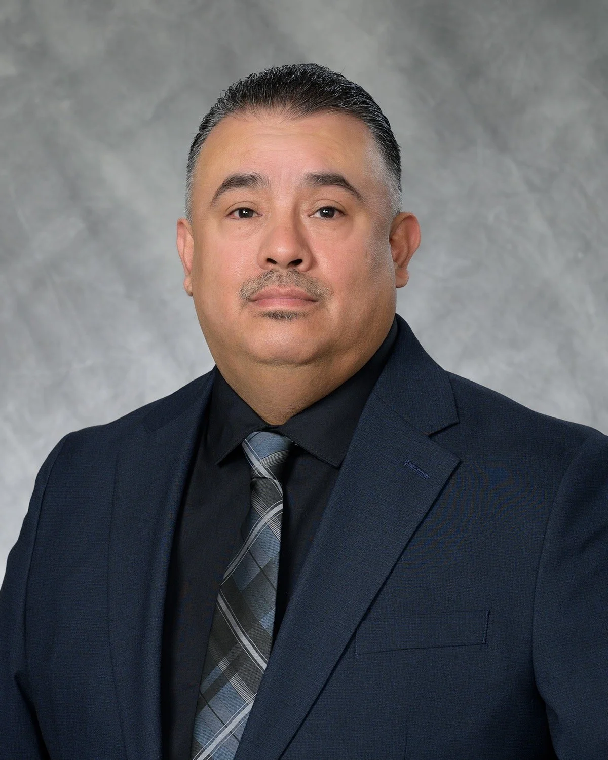 Professional headshot of a man in a dark suit, black shirt, and a plaid gray tie, with a neutral gray background.