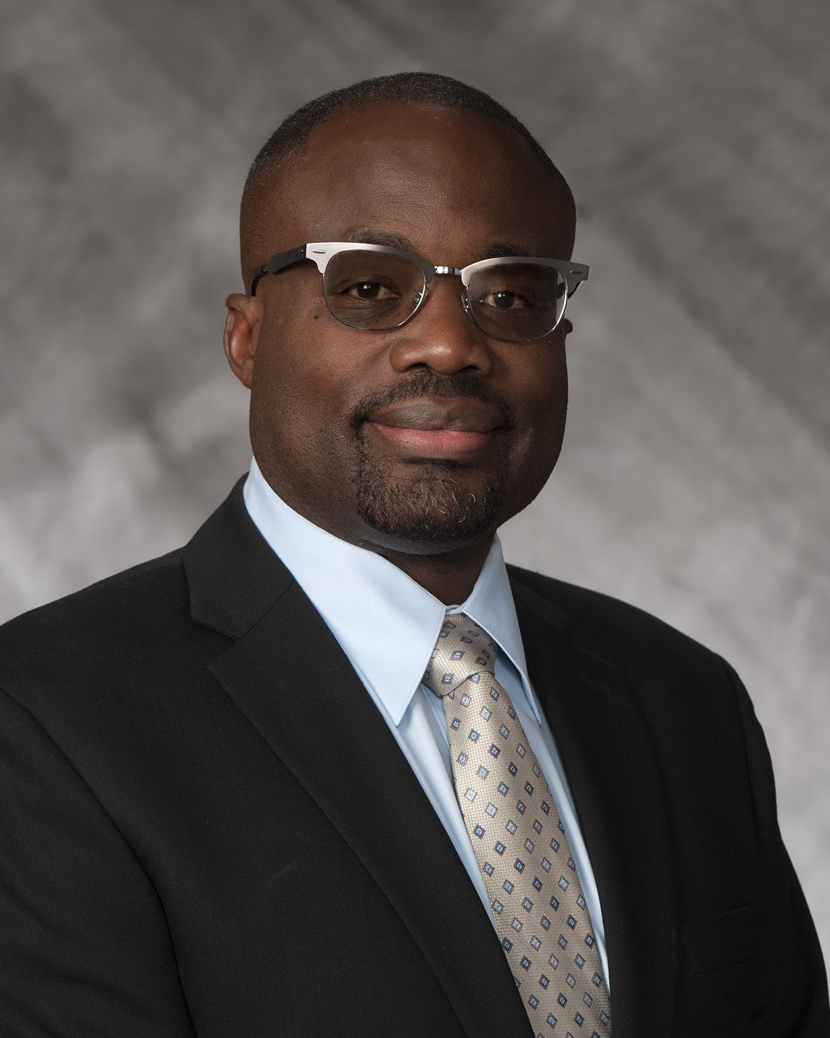 A professional man in a dark suit, light blue shirt, patterned tie, and glasses posing against a grey background.