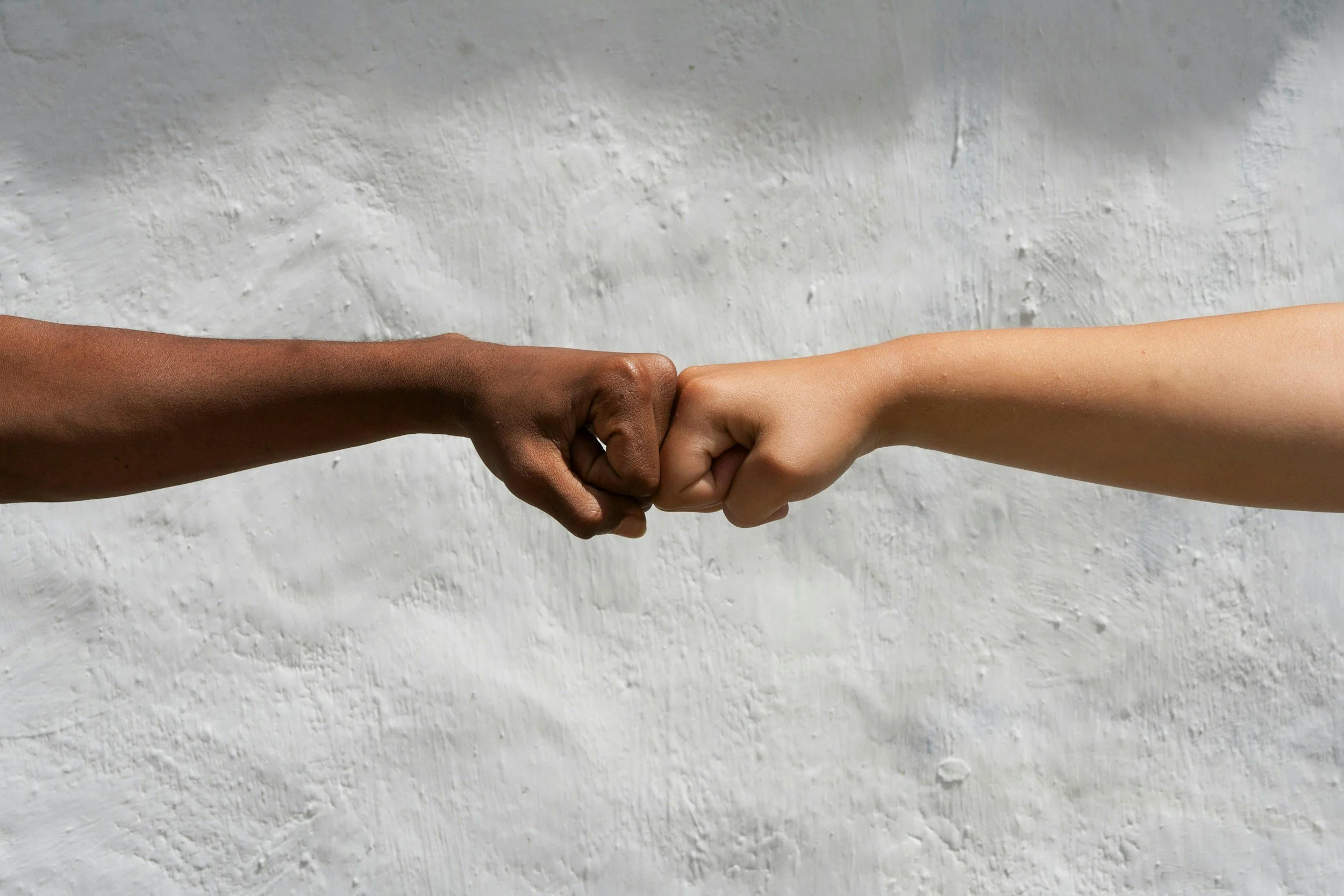 Two hands, one darker skin tone and one lighter skin tone, fist bumping against each other in front of a white textured background.