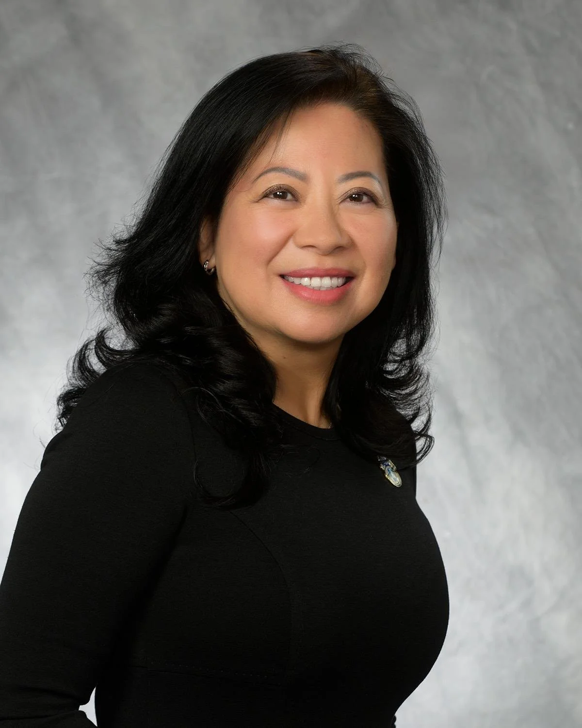 A woman with black hair smiling, wearing a black top and a pin, standing against a gray backdrop.