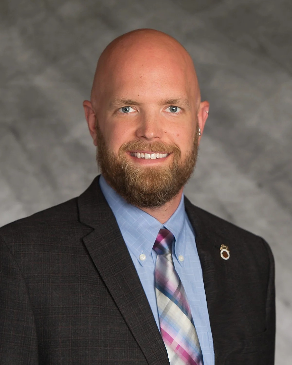Headshot of a smiling man with a bald head, beard, blue eyes, wearing a dark suit jacket, a light blue shirt, and a multi-colored striped tie, against a gray background.