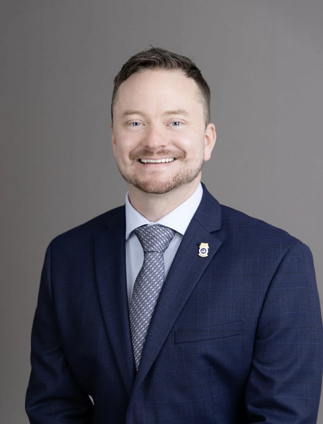 A professional headshot of a man in a navy blue suit, white shirt, and patterned tie, smiling against a gray background. He has short brown hair, a beard, and a badge pin on his lapel.