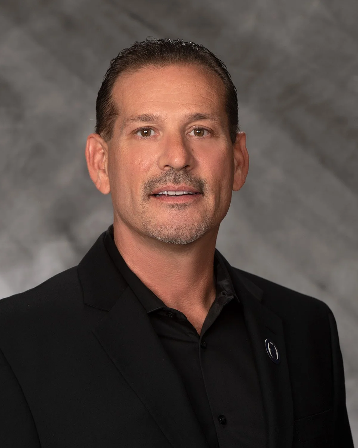 A professional headshot of a man with short dark hair, a light beard, and wearing a black suit and shirt, against a gray textured background.