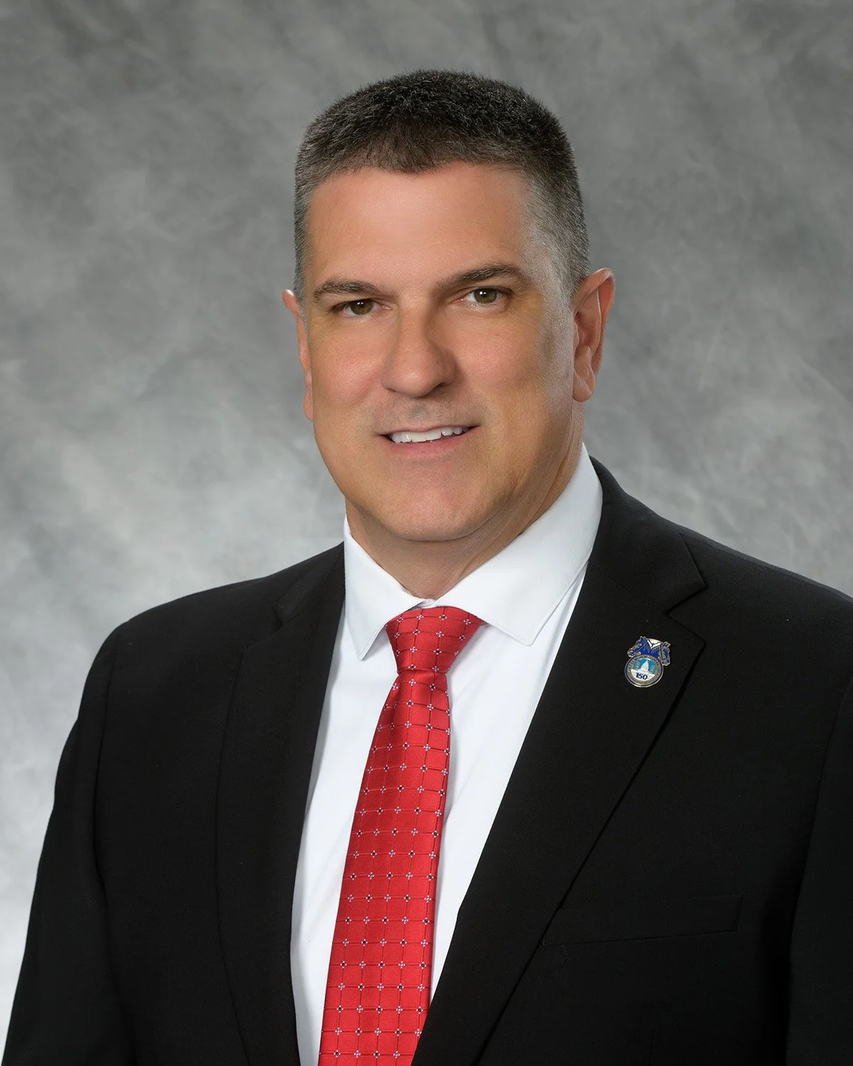 Professional headshot of a man dressed in a black suit with a white shirt and red patterned tie, gray textured background.