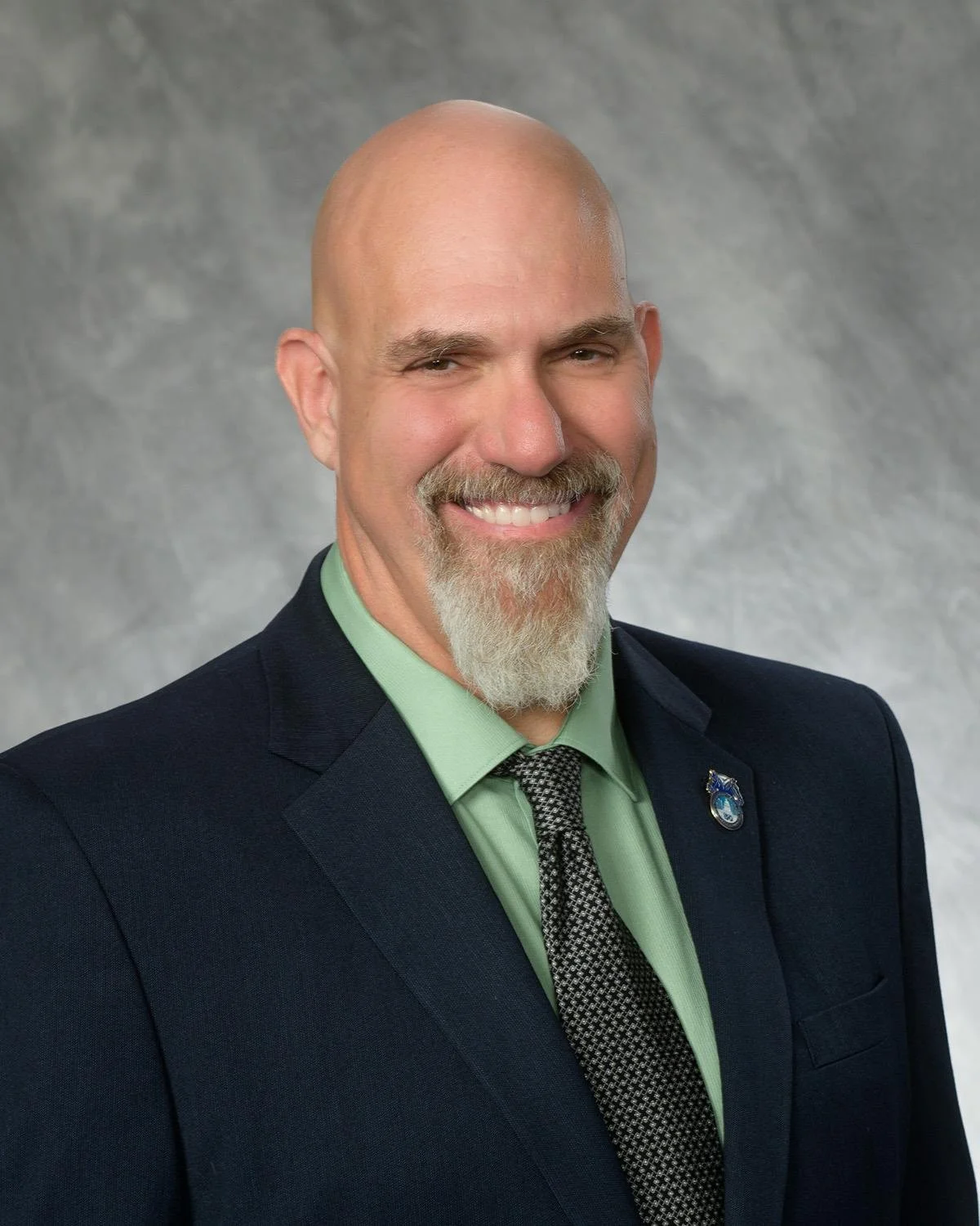 A smiling man with a bald head and gray beard dressed in a dark suit, light green shirt, and patterned tie, standing against a gray background.