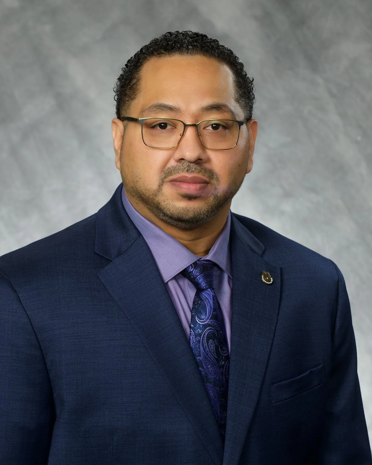 Professional headshot of a man with glasses wearing a dark blue suit, purple shirt, and a purple patterned tie, standing against a gray background.