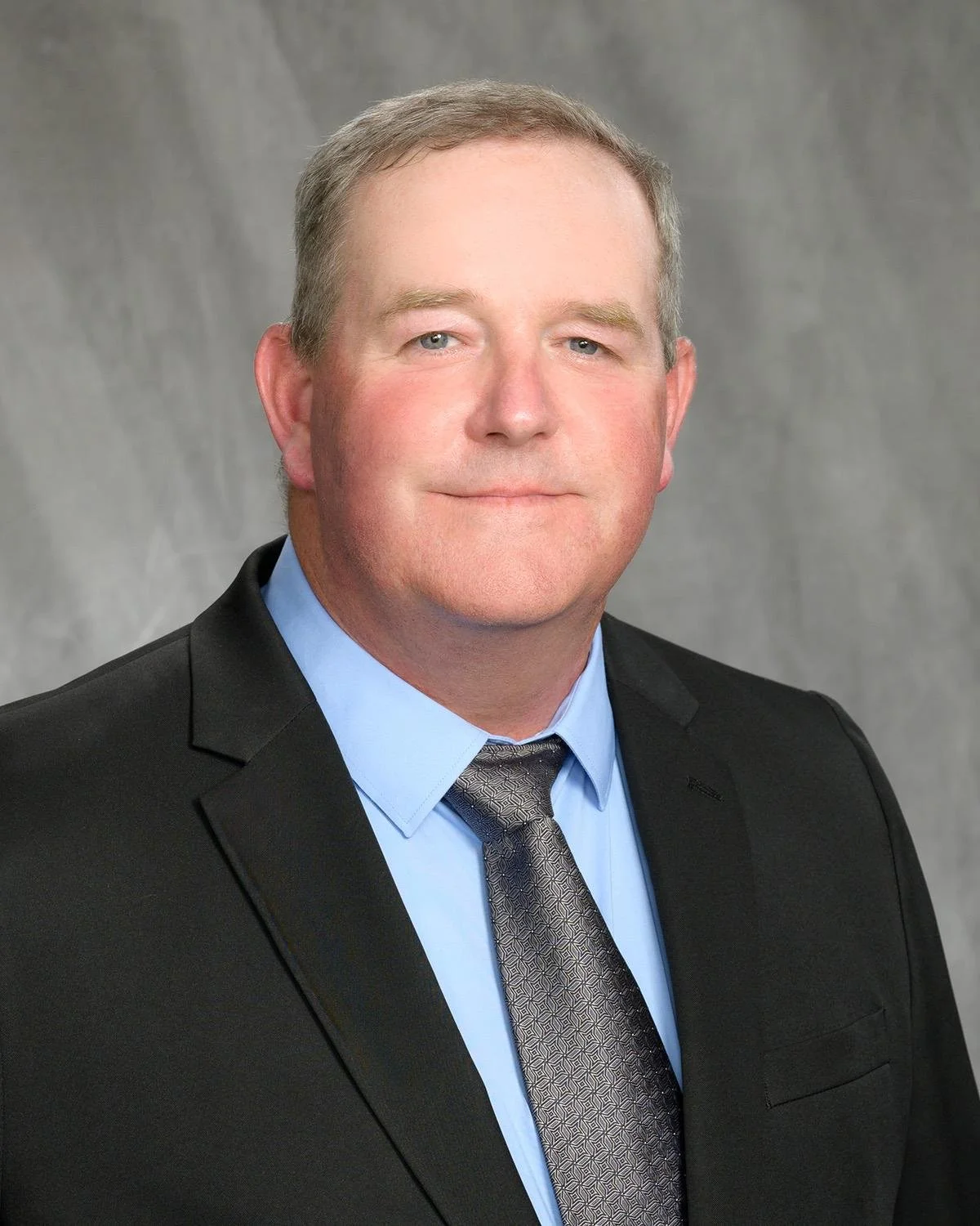 Professional headshot of a man wearing a black suit, light blue dress shirt, and a patterned gray tie, against a gray background.