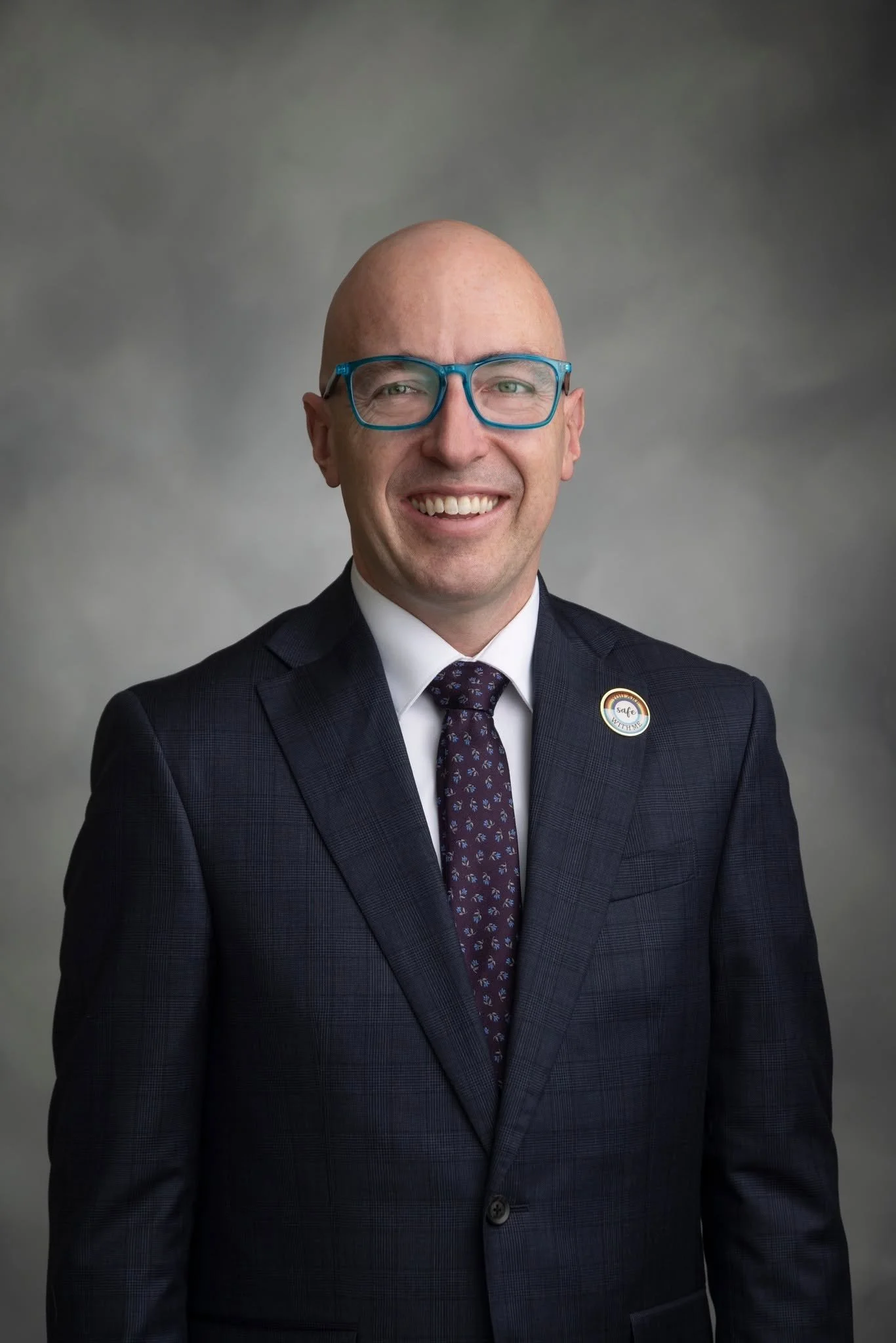A man with a bald head and wearing bright blue glasses, a dark plaid suit, white shirt, and patterned tie, smiling at the camera against a gray studio background.