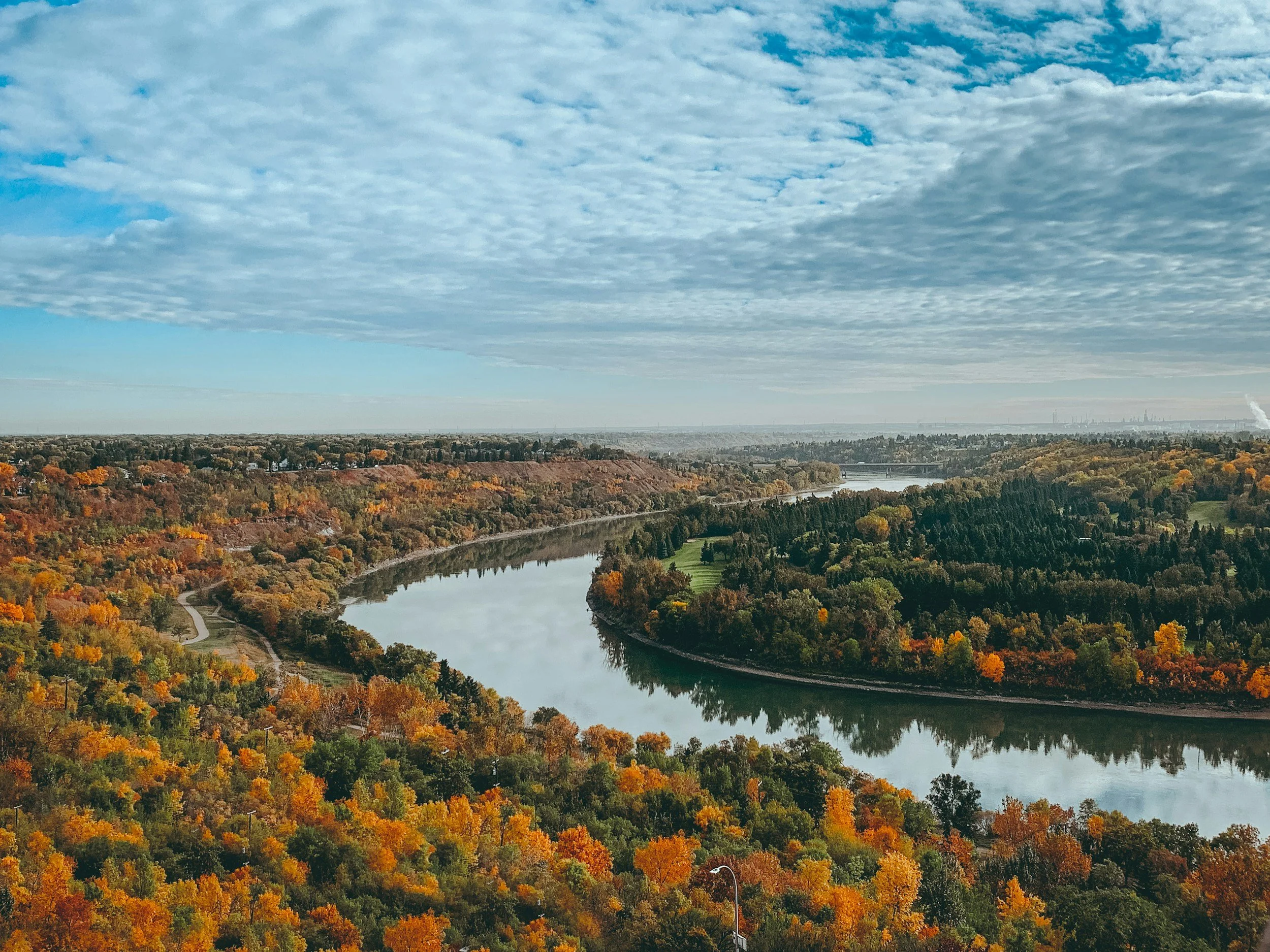 Aerial view of a river winding through forested hills during autumn, with colorful fall foliage and a partly cloudy sky.