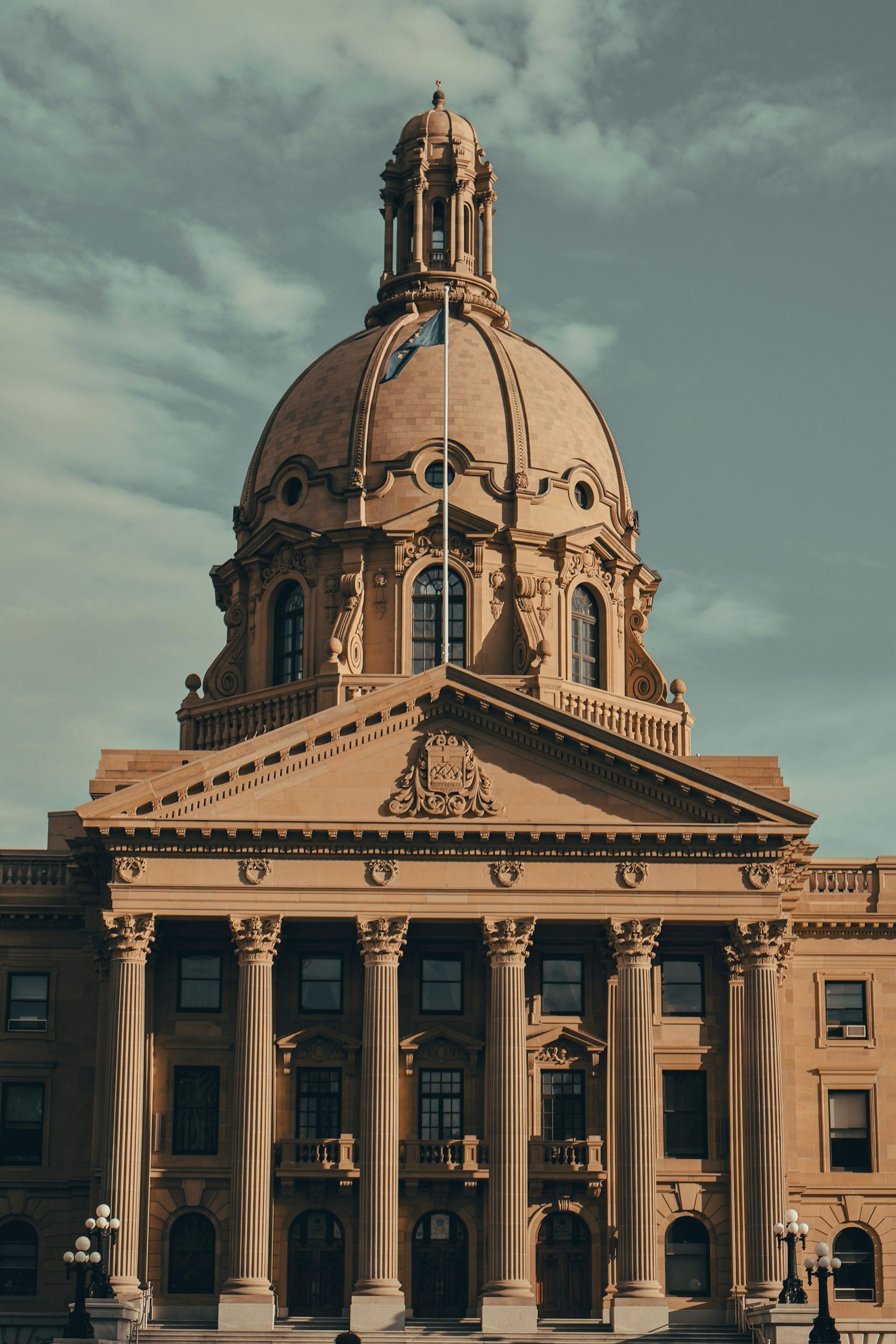 The exterior of a historic government building with classical architecture, featuring tall columns, ornate details, and a large dome on top, under a partly cloudy sky.