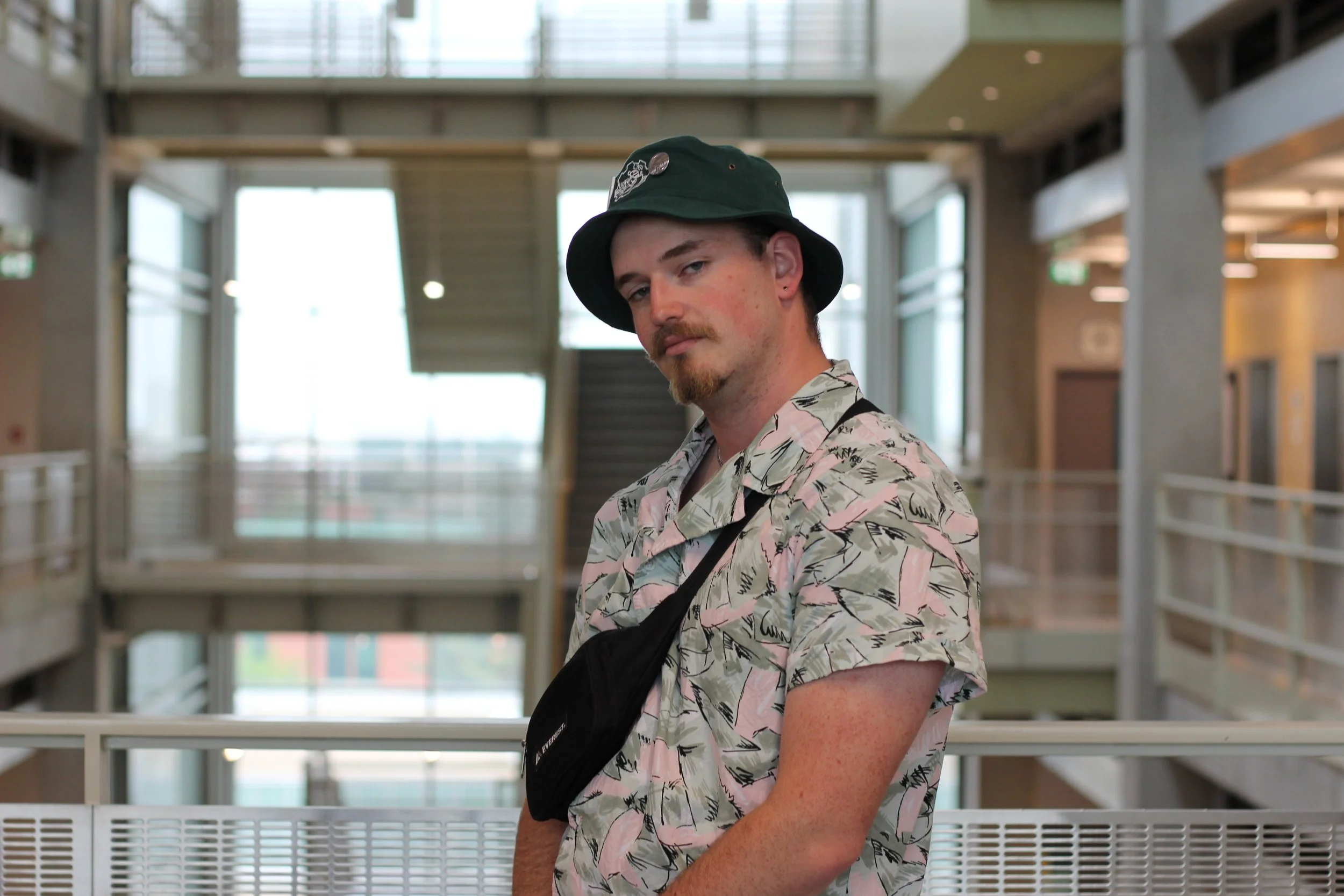 A young man with a beard and mustache wearing a patterned short-sleeve shirt, black bucket hat, and a black crossbody bag, standing inside a modern, multi-level building with open walkways and glass walls.