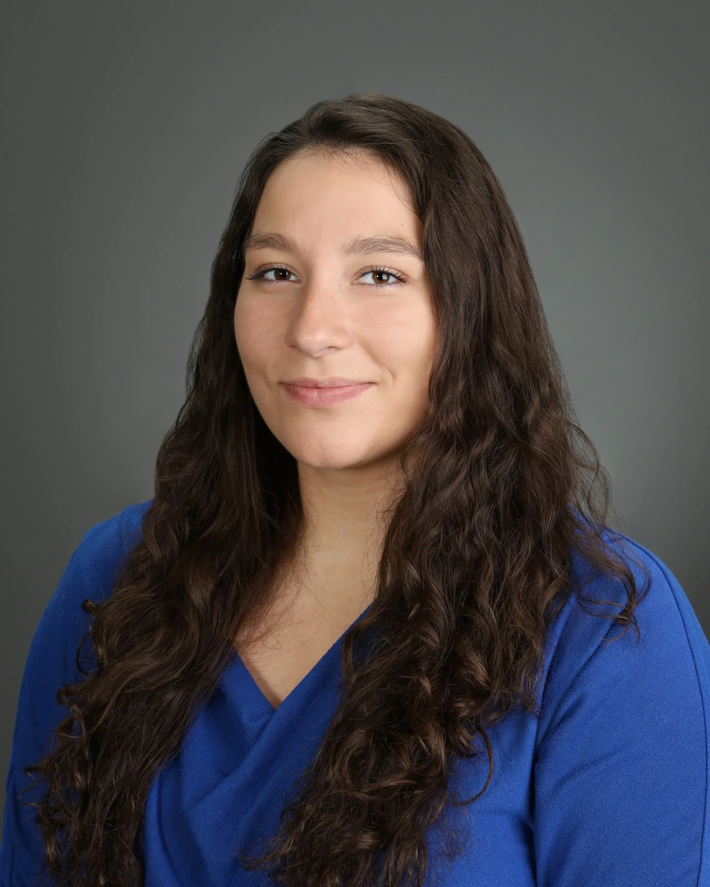 Head and shoulders portrait of a young woman with long, wavy brown hair, wearing a blue shirt, smiling softly against a gray background.