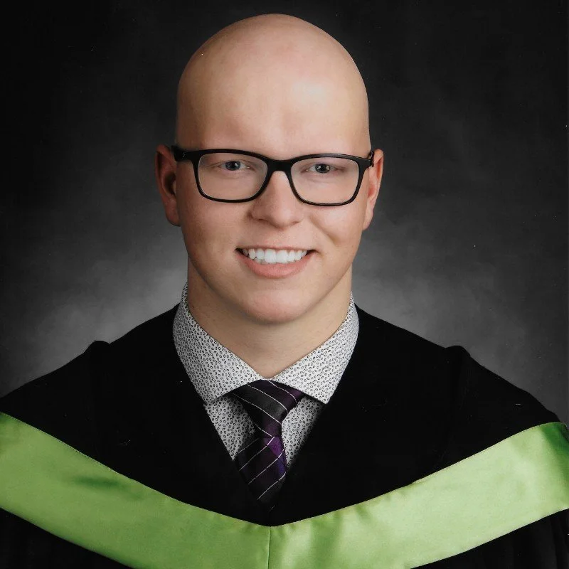 A young man in graduation attire, wearing black glasses, a black gown with green accents, a white shirt with small black dots, and a striped tie, smiling against a dark background.