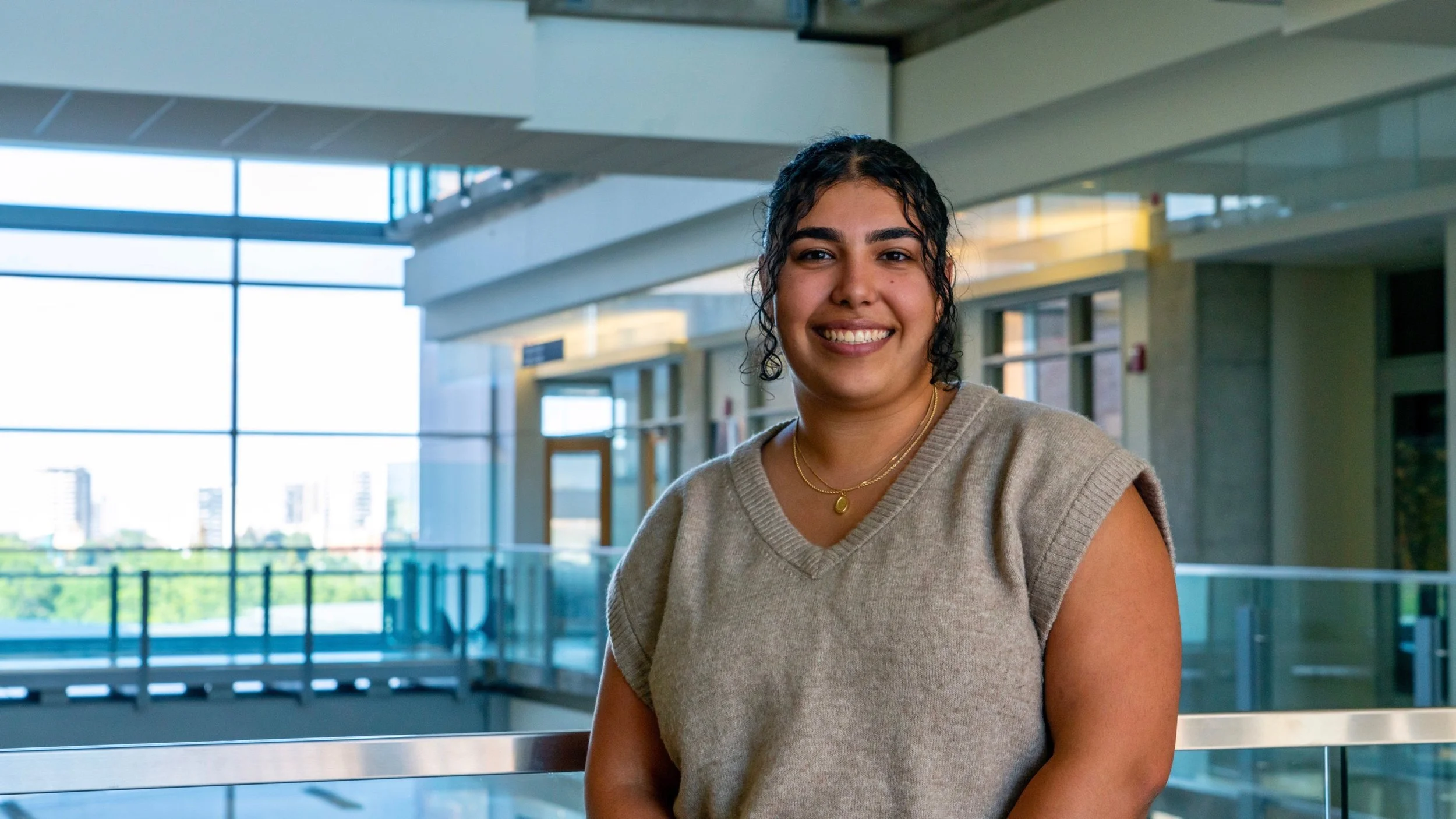 A smiling woman with dark curly hair wearing a beige sleeveless sweater and layered gold necklaces standing in a modern building with large windows and an indoor balcony.