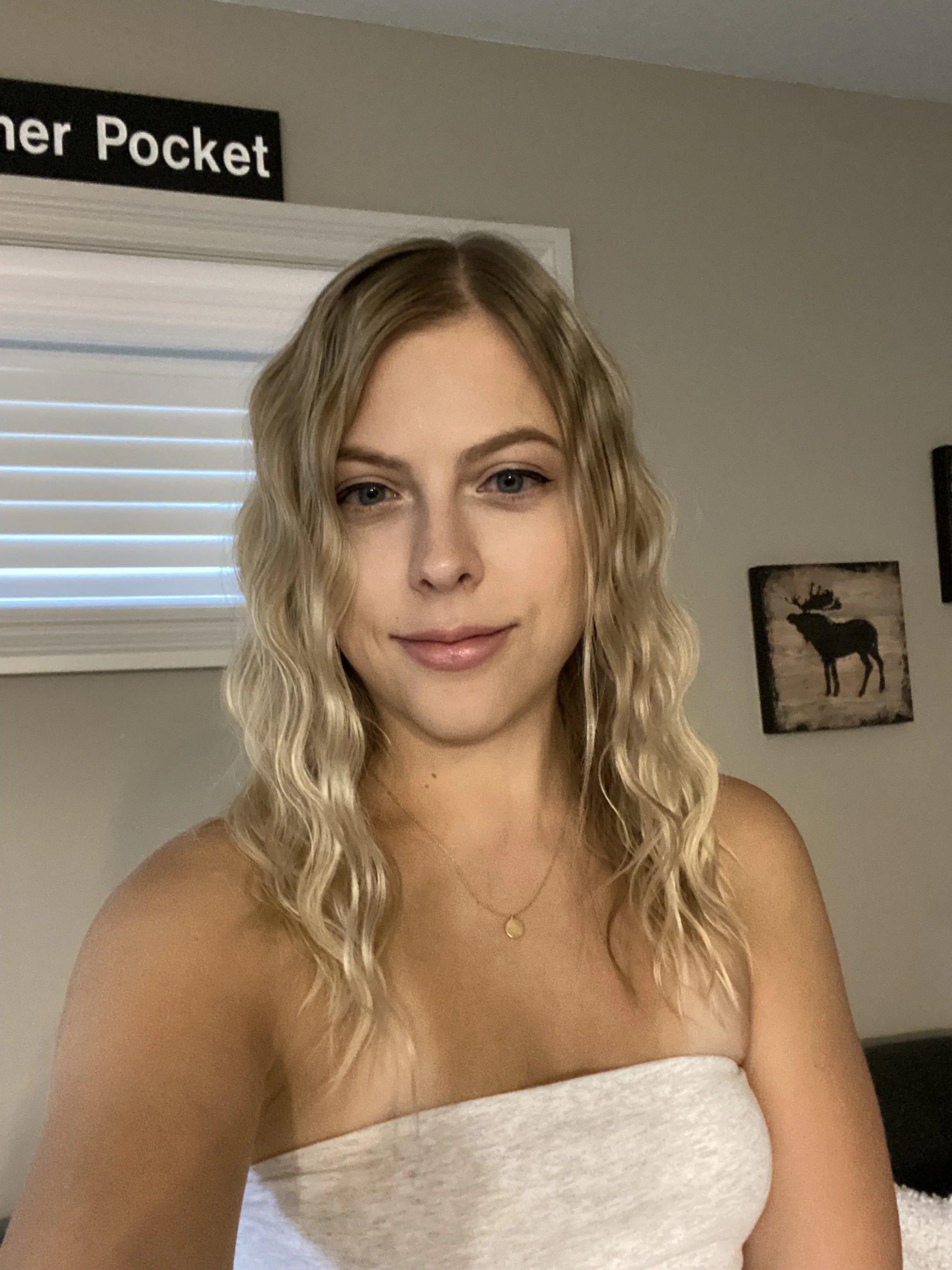 A young woman with wavy blonde hair taking a selfie indoors, wearing a strapless white top and a gold necklace.