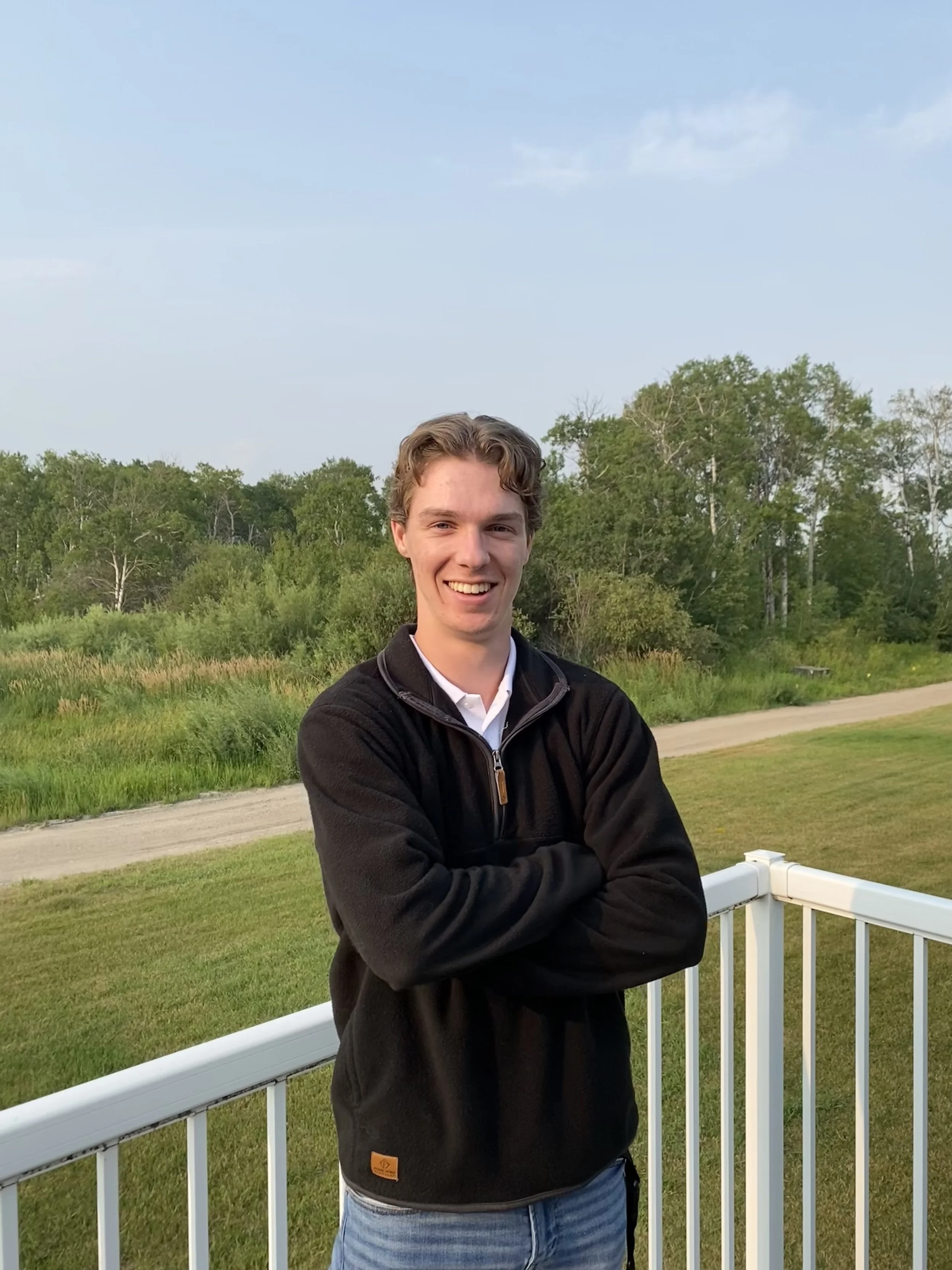 A young man with light brown hair, wearing a black zip-up jacket and blue jeans, smiling with arms crossed, standing outdoors on a balcony with a grassy field, trees, and a blue sky in the background.