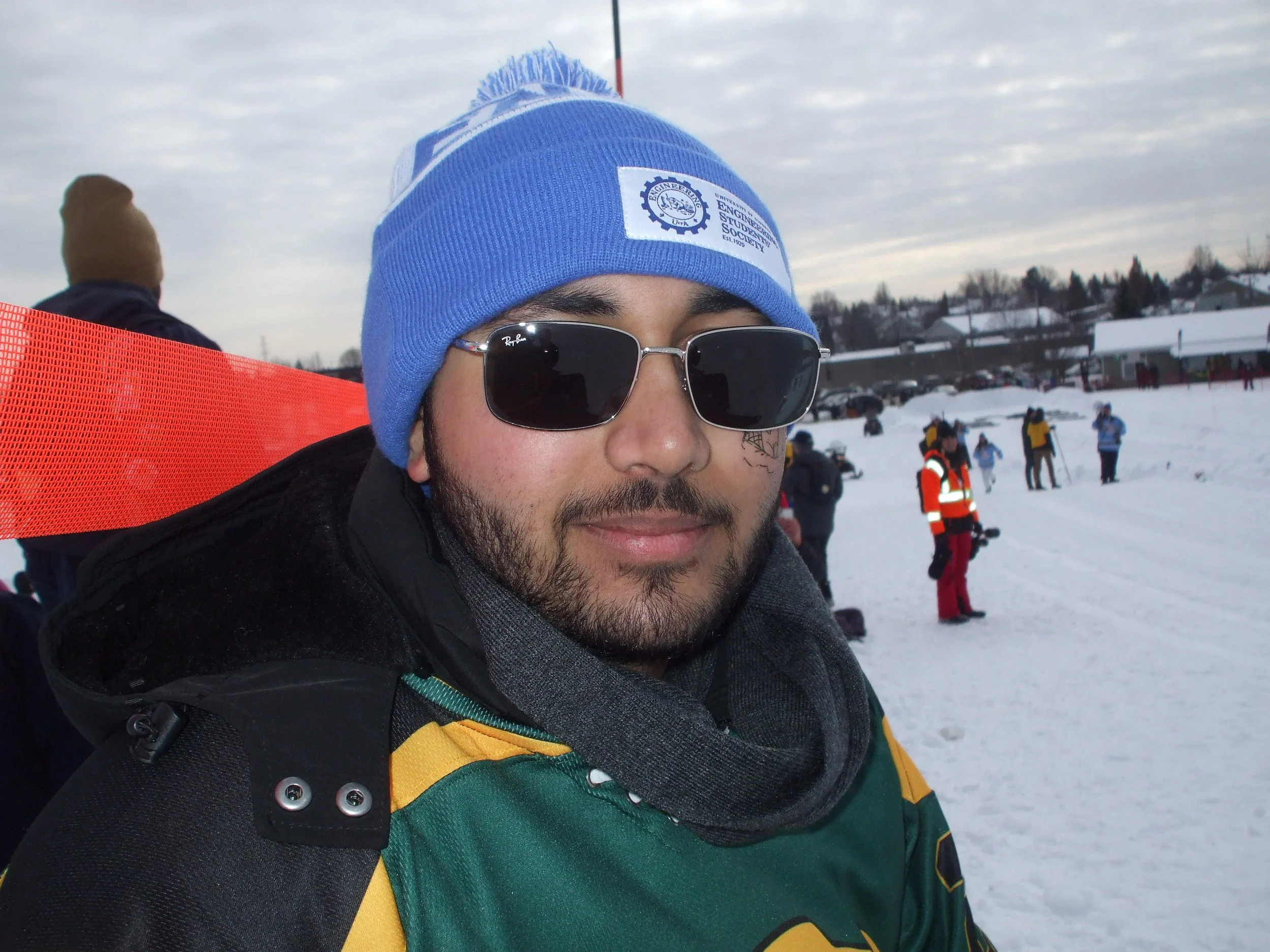 Man wearing sunglasses, a blue beanie with a logo, and a green and yellow jacket, standing in a snowy outdoor setting with other people in the background.