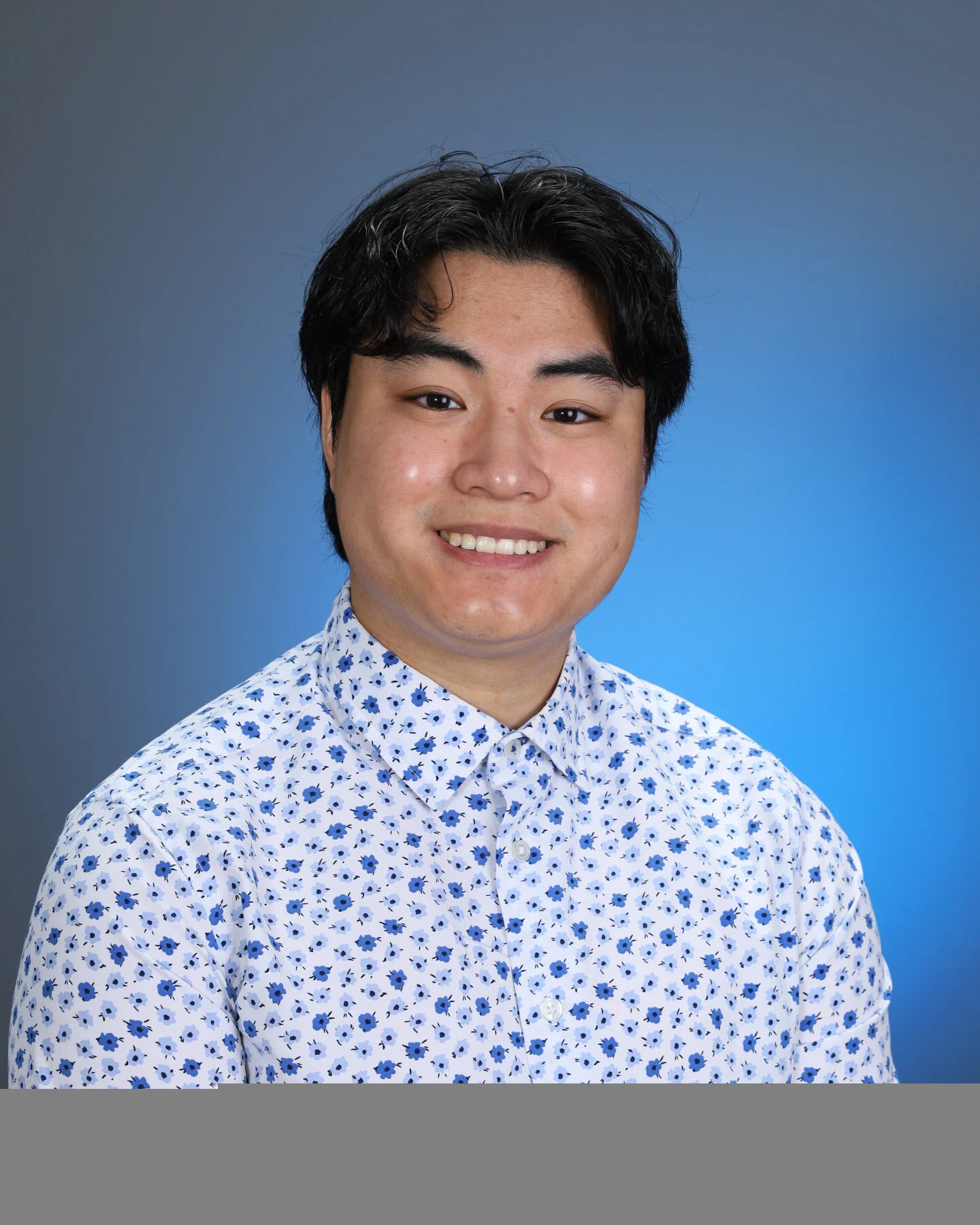 Young man with black hair smiling, wearing a white shirt with blue floral pattern, against a gradient blue background.