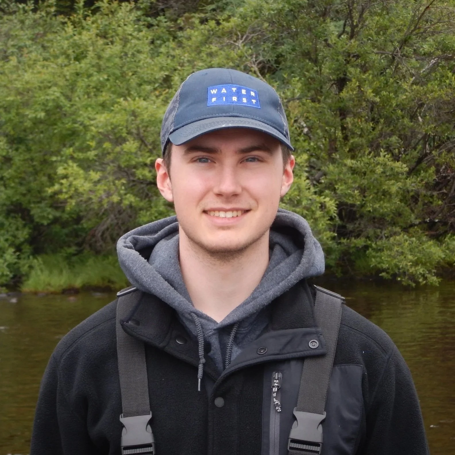 A young man smiling outdoors in front of greenery and a body of water, wearing a baseball cap with 'Water First' on it, a hoodie, and a black jacket.