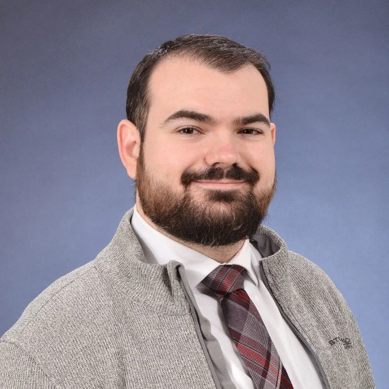 A man with dark hair and a beard, wearing a gray sweater over a white collared shirt and a striped tie, smiling in front of a blue gradient background.
