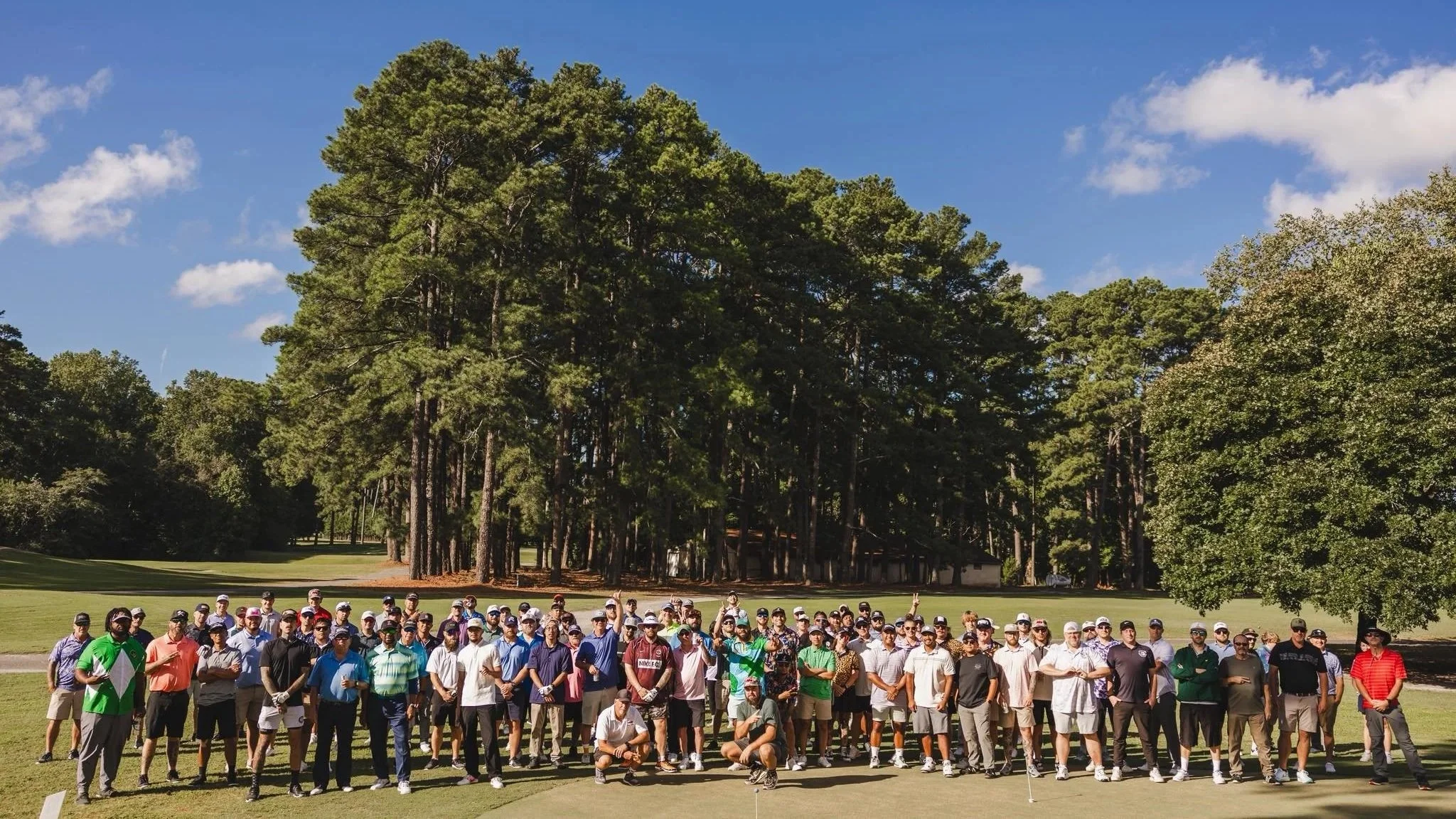 Group of people gathered outdoors on a golf course with tall trees and a clear blue sky in the background.