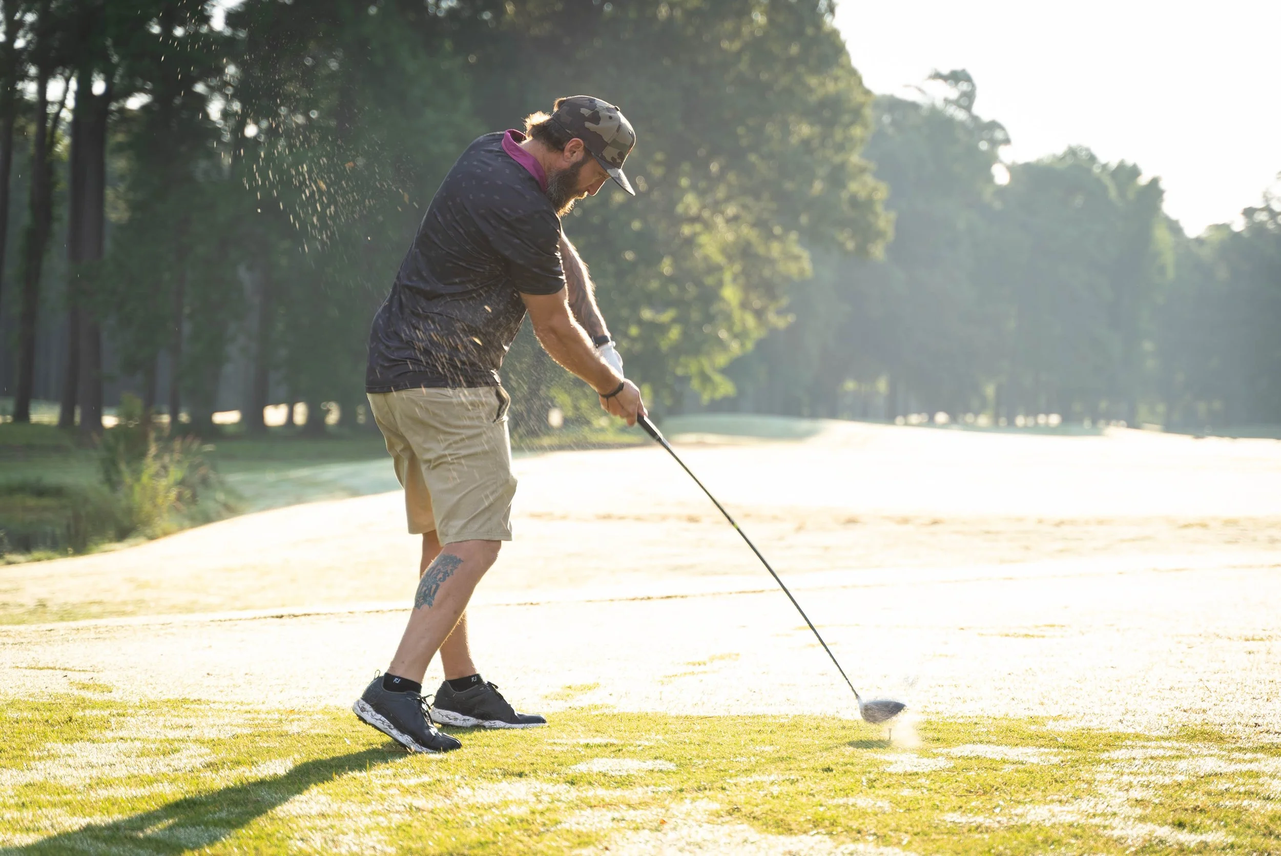 A man wearing a camouflage hat, dark shirt, and khaki shorts is playing golf on a sunny day, preparing to hit a golf ball on a grassy course with trees in the background.