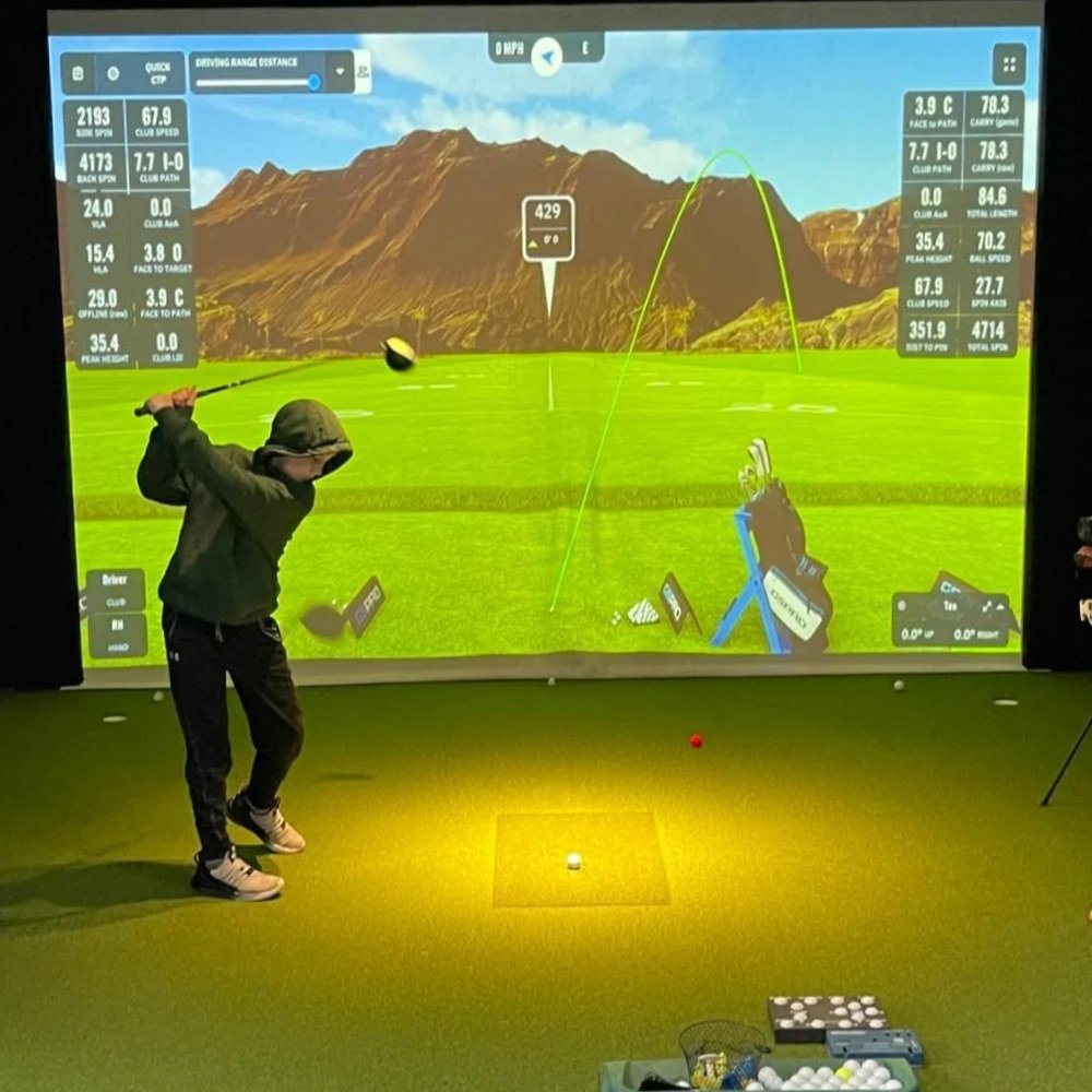 A person practicing golf in an indoor golf simulator, swinging a golf club at a projected screen showing a golf course with mountains in the background and various golf statistics.