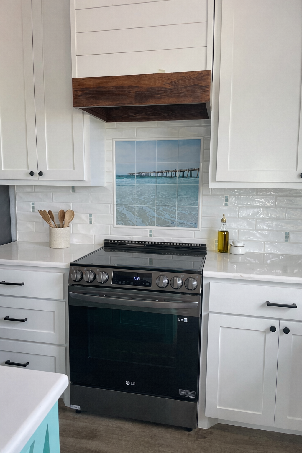 Kitchen with white cabinets, a black stove, a picture of a pier over water on the wall, and utensils in a white container.
