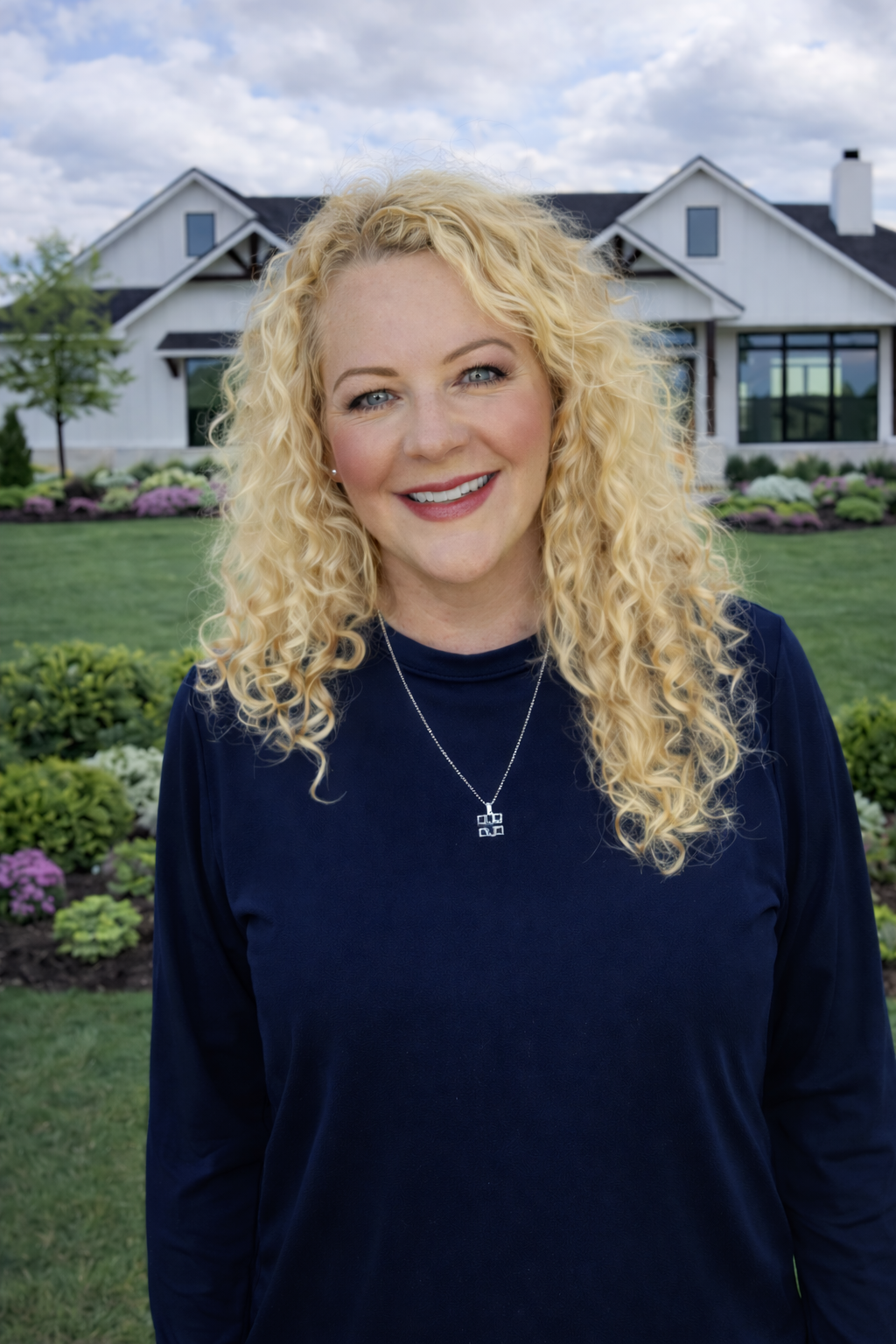 A woman with curly blonde hair smiling outdoors in front of a house and a landscaped garden.