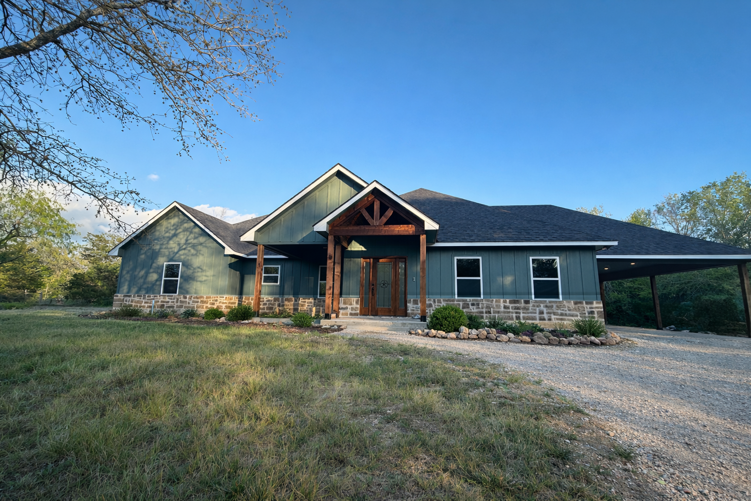 A modern single-story house with dark green exterior walls, stone accents at the base, and a gabled roof, surrounded by a grassy lawn and trees under a blue sky.