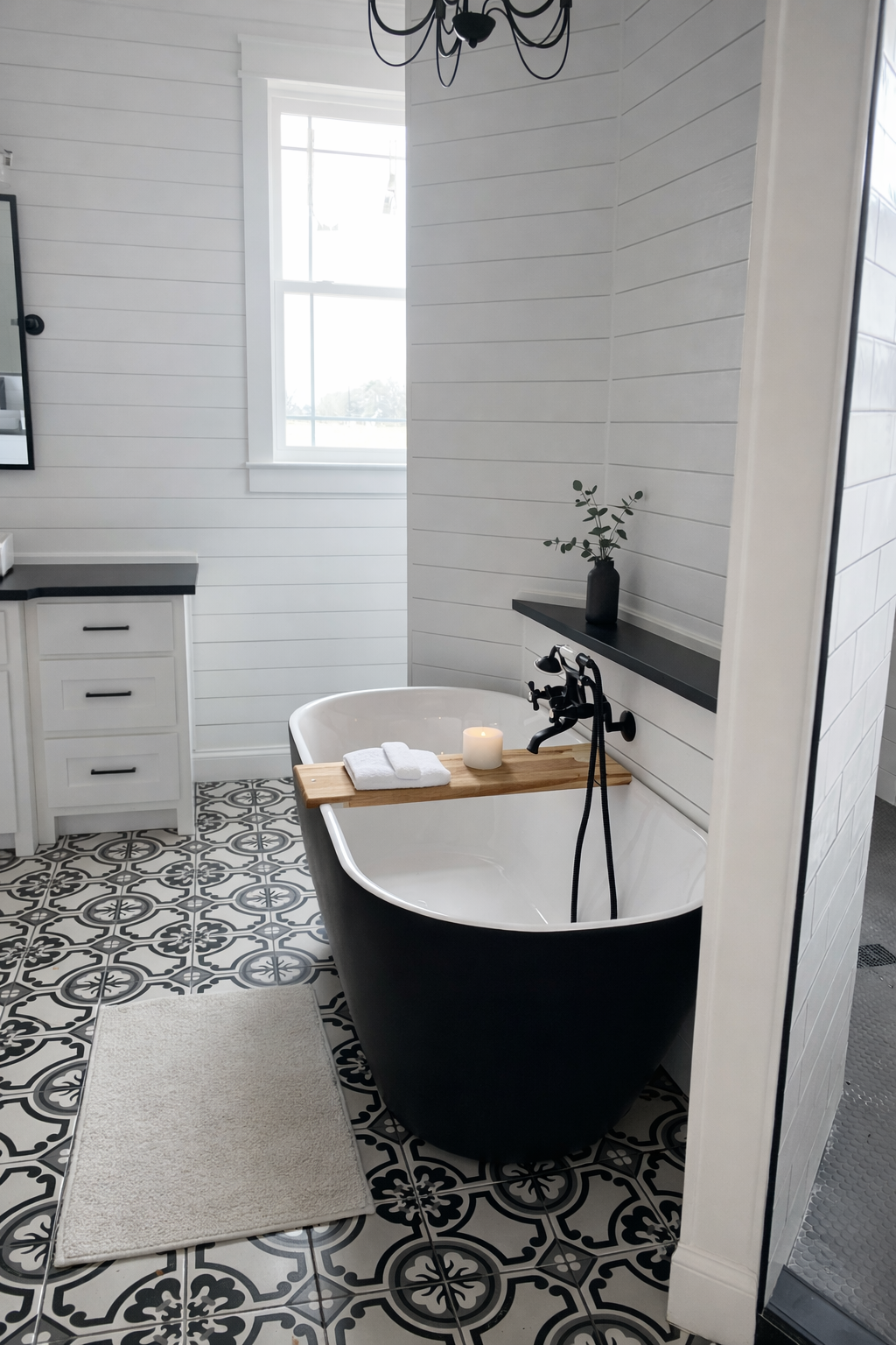 Bathroom with black clawfoot tub featuring a wooden tray, candle, and towels. White shiplap walls, black and white patterned floor, window, and black accents.
