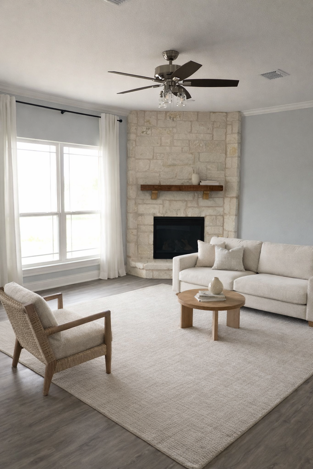 Living room with a beige sofa, wooden coffee table, armchair, stone fireplace with wooden mantel, large window with white curtains, ceiling fan, and light gray walls.